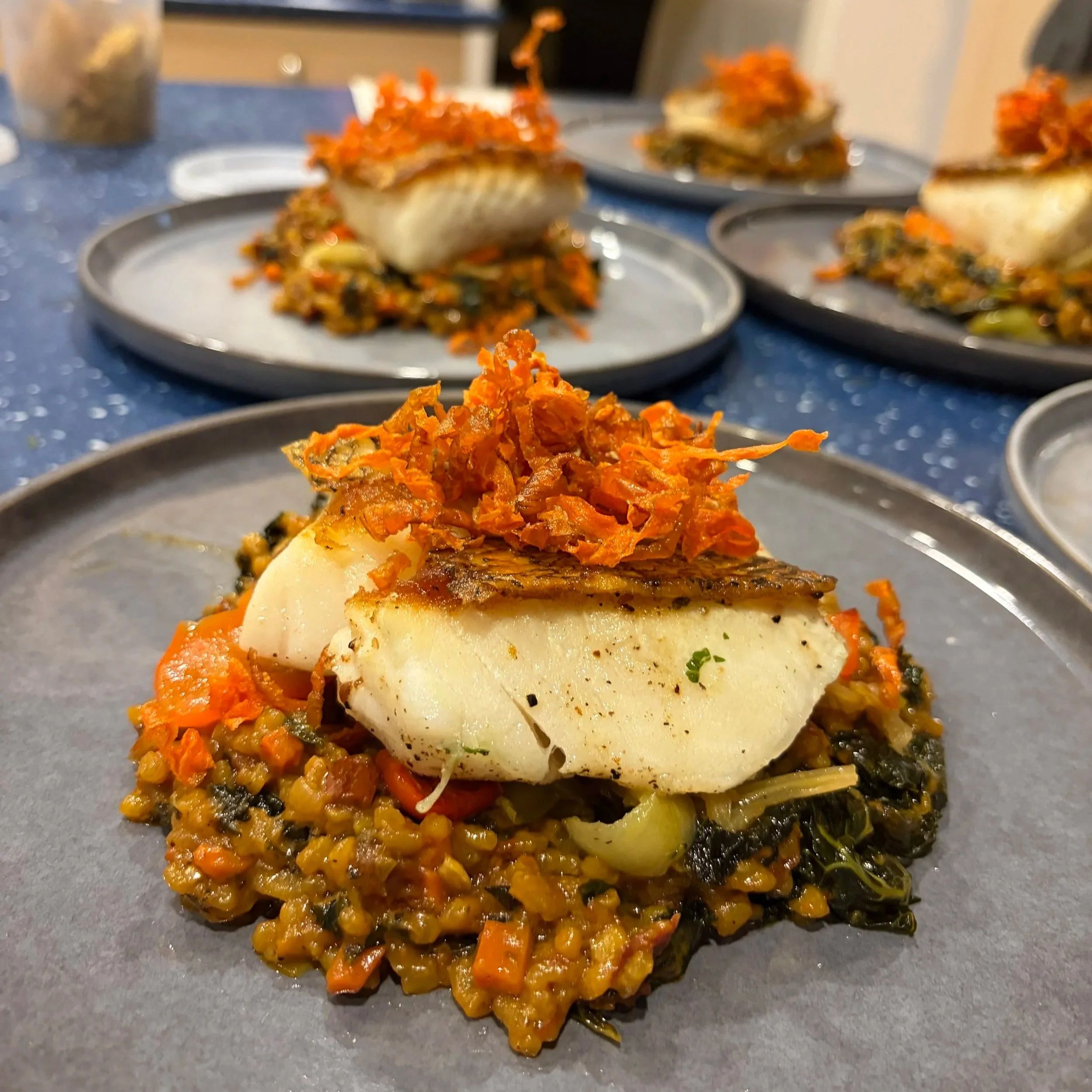 Close-up of a plated dish featuring a piece of fish topped with shredded carrots and herbs, served over a bed of cooked lentils and vegetables, on a dark plate.