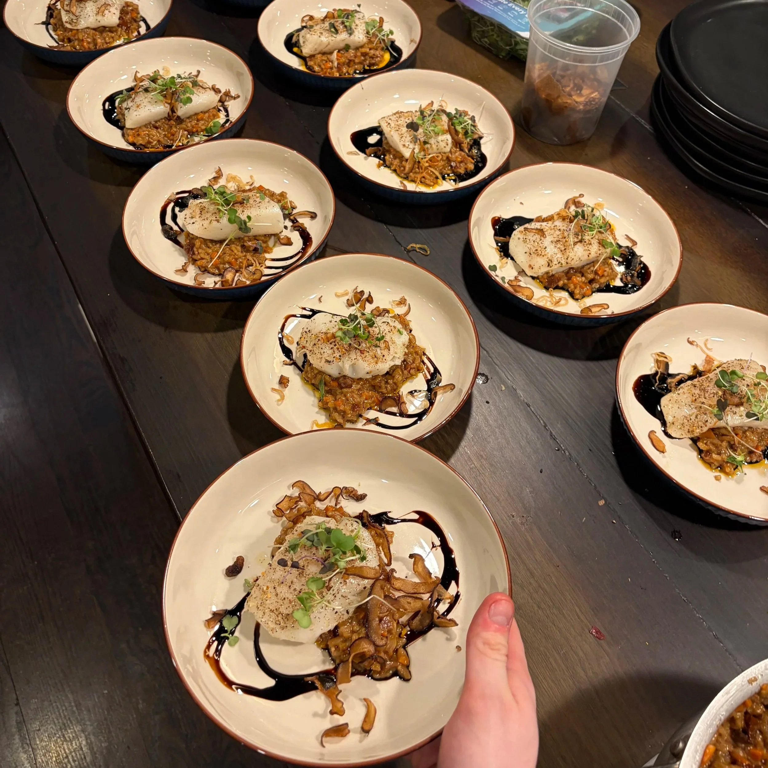 Multiple small white bowls filled with a gourmet dish consisting of a white fish fillet served on a bed of seasoned rice and vegetables. Each dish is garnished with microgreens and drizzled with balsamic glaze, with some dishes also topped with crisp