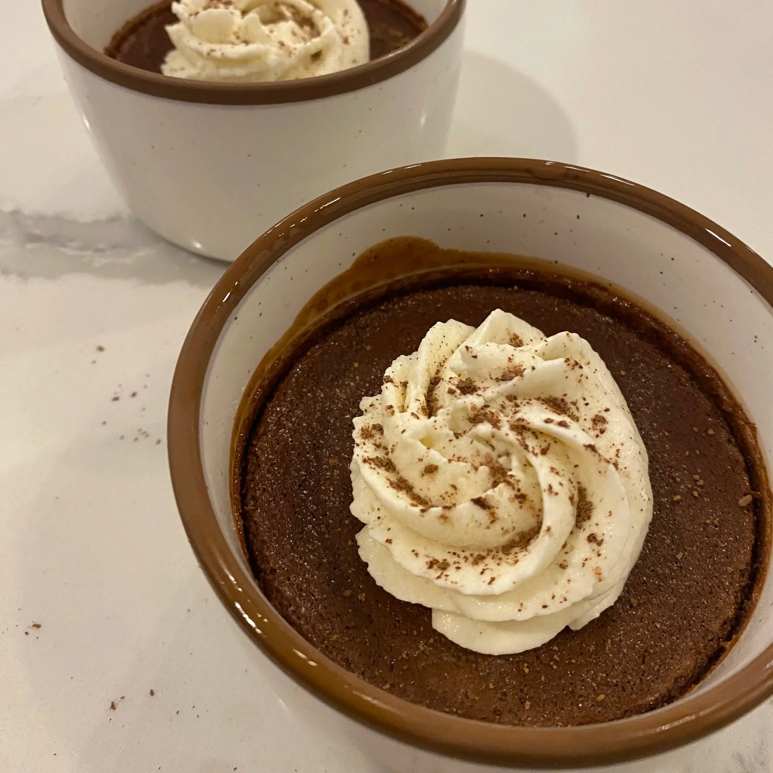 A close-up of a small bowl of chocolate dessert topped with whipped cream and a sprinkle of cocoa or cinnamon powder, with a second similar bowl blurred in the background.