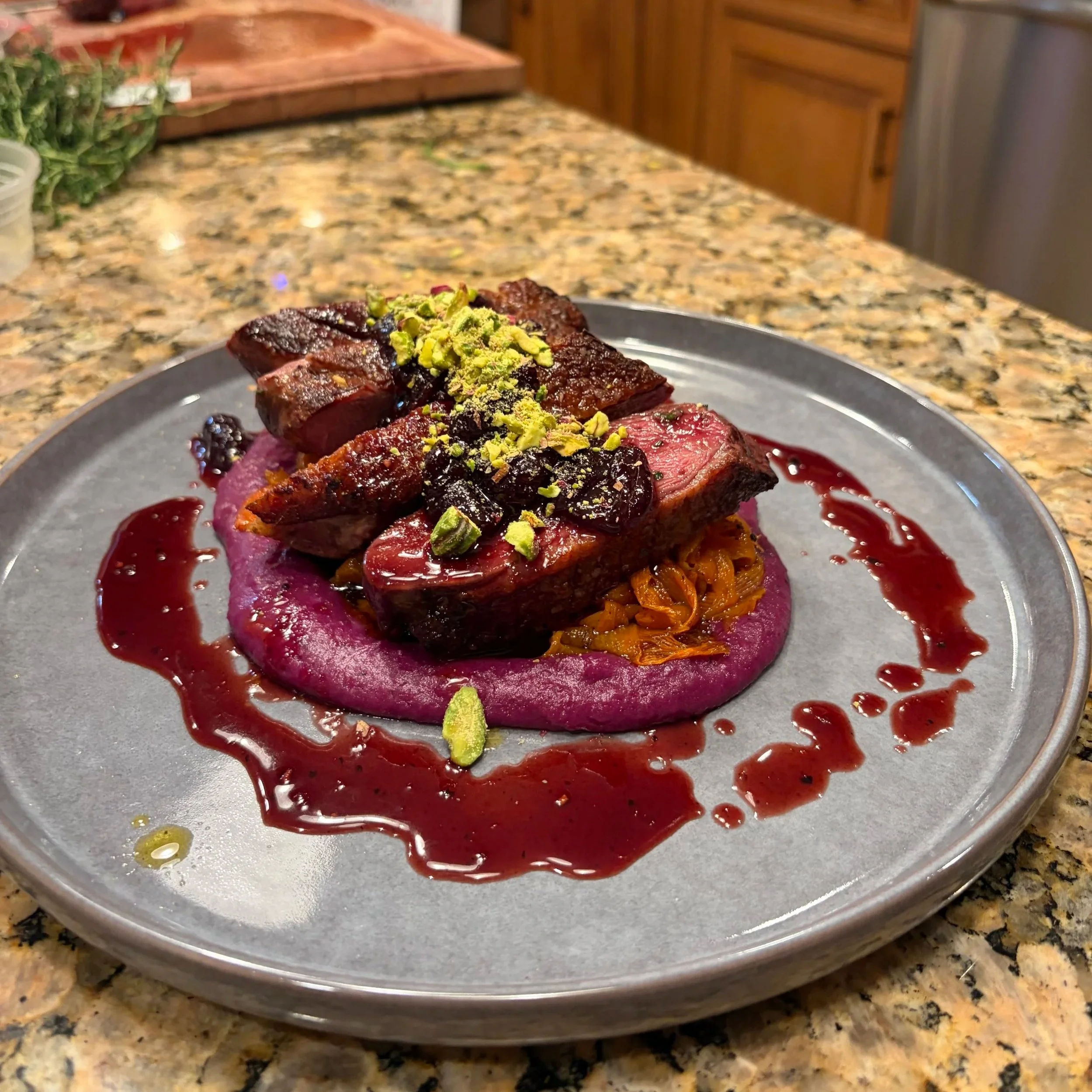 A plated dish featuring a piece of cooked meat topped with chopped pistachios, surrounded by purple sauce and garnished with blueberries and orange slices on a gray plate set on a speckled granite countertop.