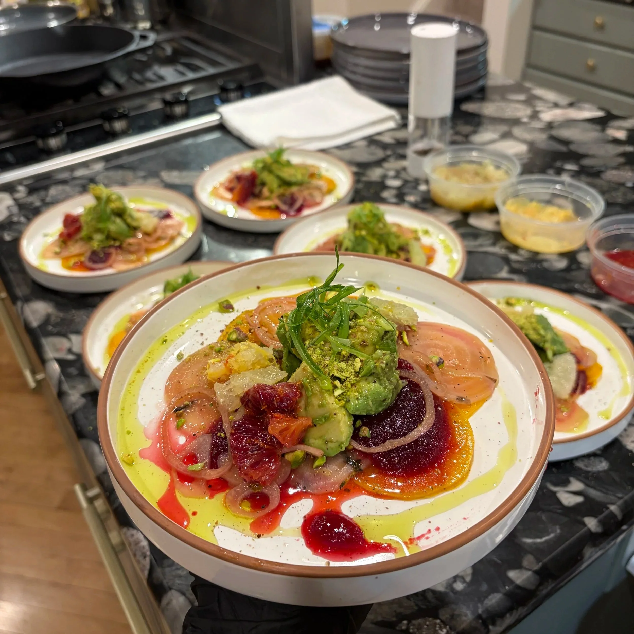 Close-up of several plates with colorful food dishes, garnished with vegetables, on a kitchen counter with plates, containers, and a paper towel in the background.