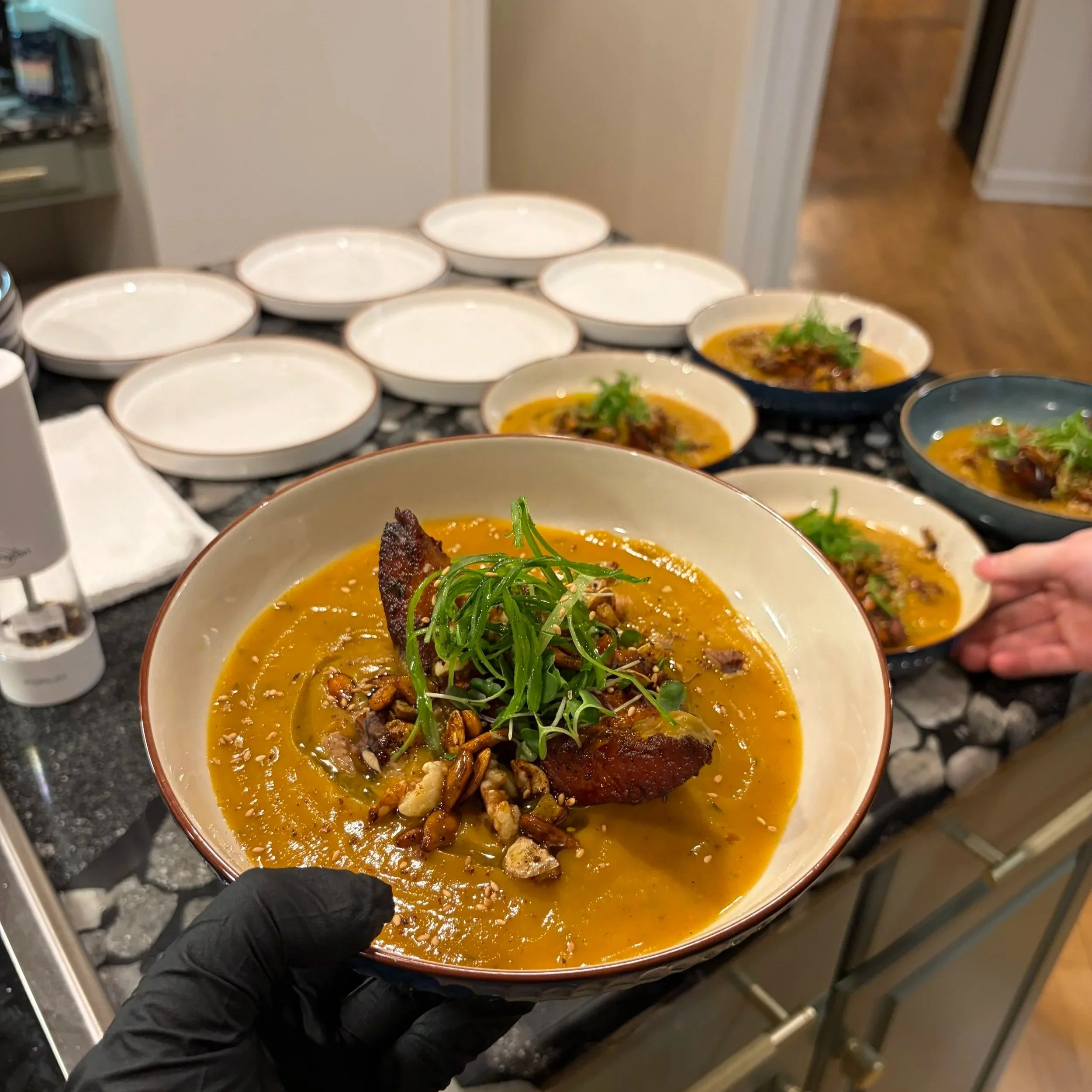 A hand holding a bowl of ramen with broth, green garnish, and meat, with multiple empty bowls in the background.