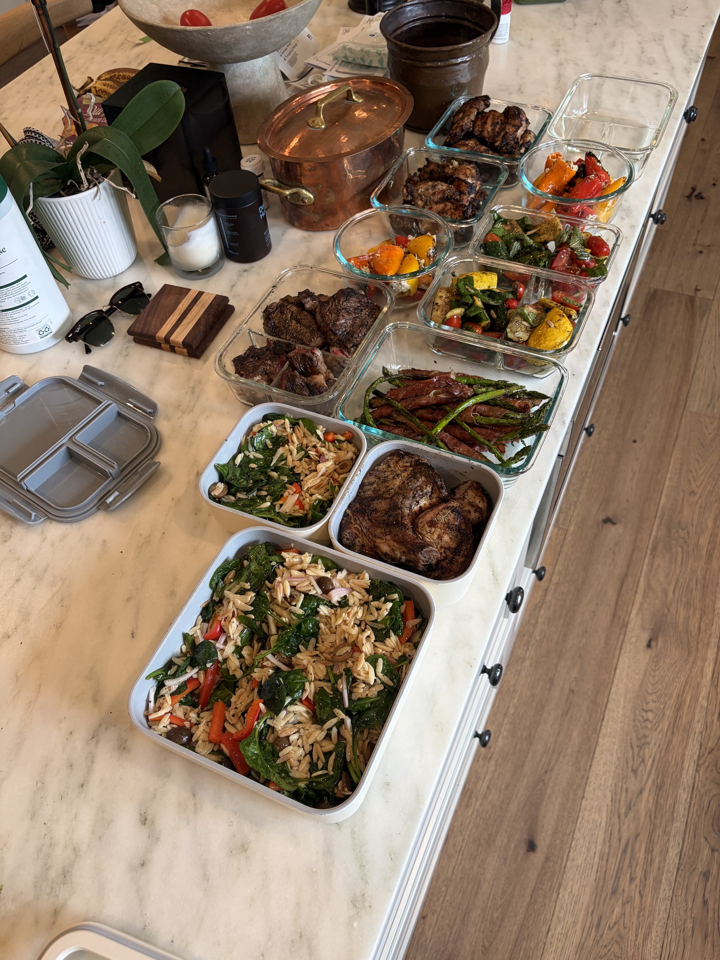 A marble kitchen counter with various dishes in glass and plastic containers, including roasted meats, grilled vegetables, pasta salad with vegetables, and cooked green beans wrapped in bacon, along with a potted orchid, eyeglasses, a coaster, and kitchen items.