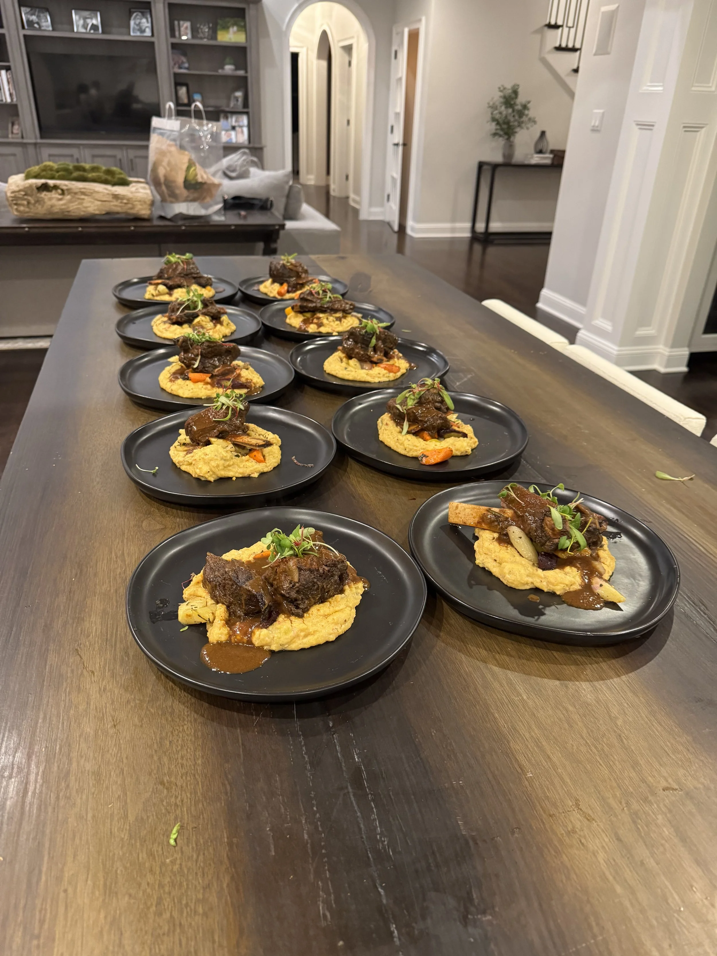 Multiple black plates with beef stew on top of mashed potatoes arranged on a wooden dining table in a living room setting with a TV, shelving, and stairs in the background.