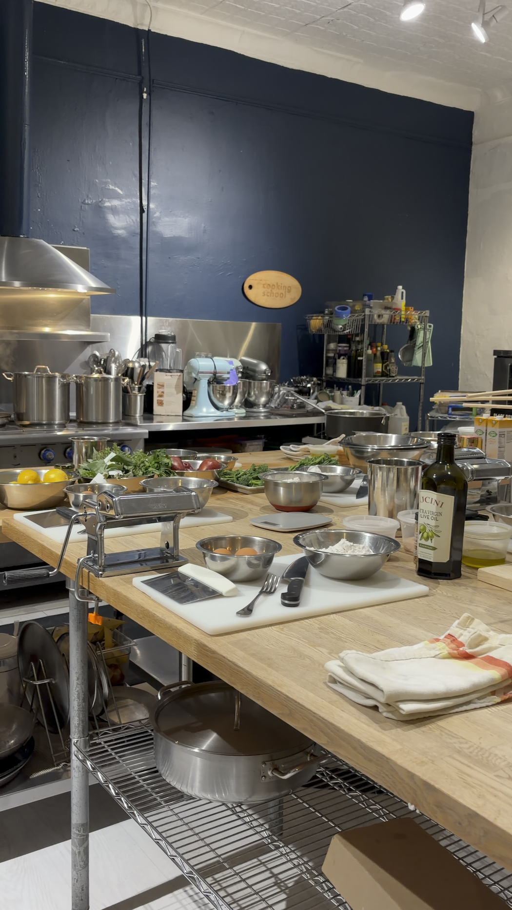 A professional kitchen with a large wooden work table covered with various ingredients, utensils, and cooking equipment. There are bowls of vegetables, measuring scale, a pasta machine, bottles of oil, and other cooking supplies. The background has shelves stocked with spices and ingredients, a blue wall with a small wooden sign that reads 'cooking school,' and overhead lights illuminating the workspace.