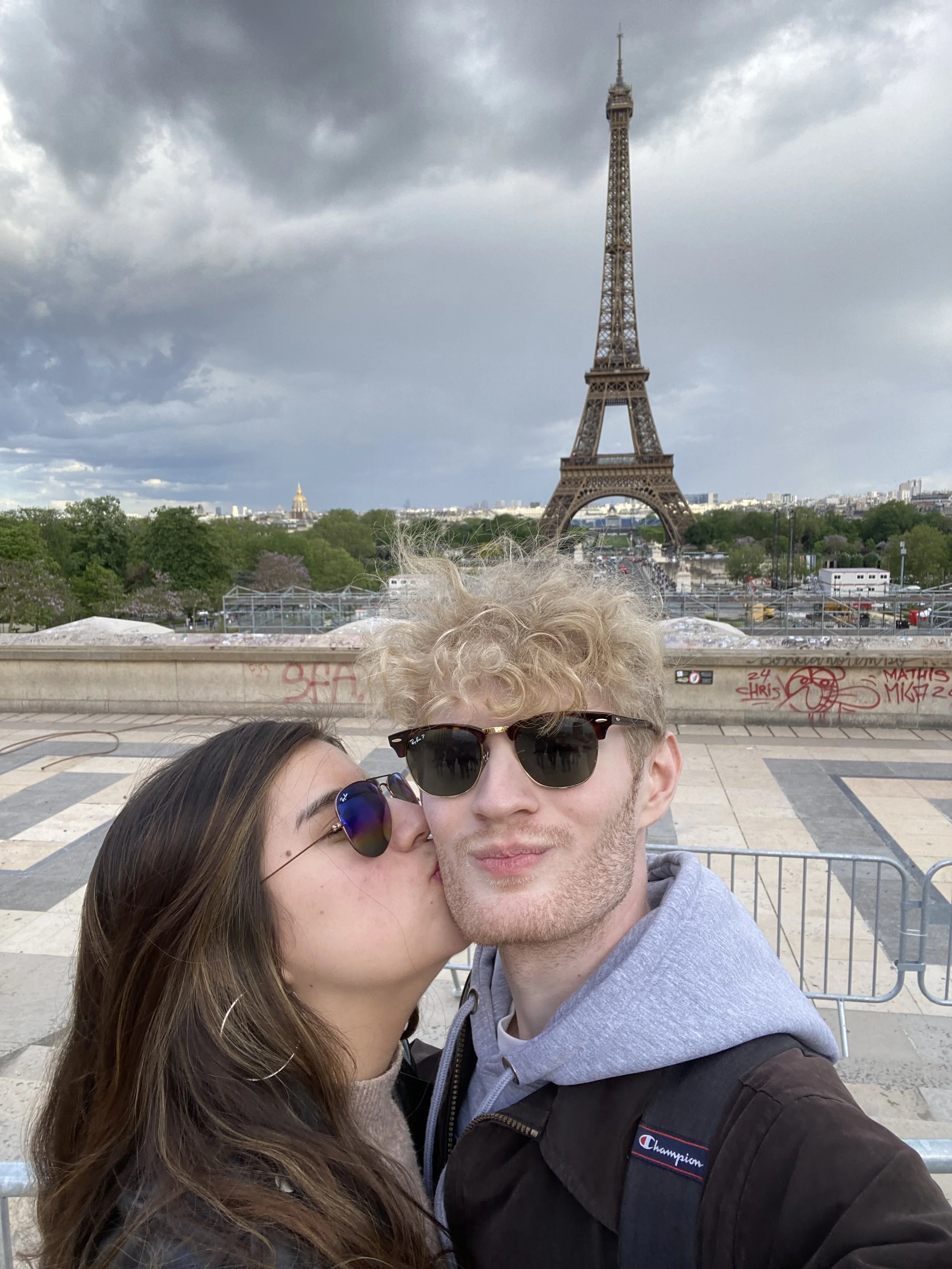 A young woman with long brown hair and sunglasses kissing a young man with curly blonde hair and sunglasses, with the Eiffel Tower in the background under cloudy skies.