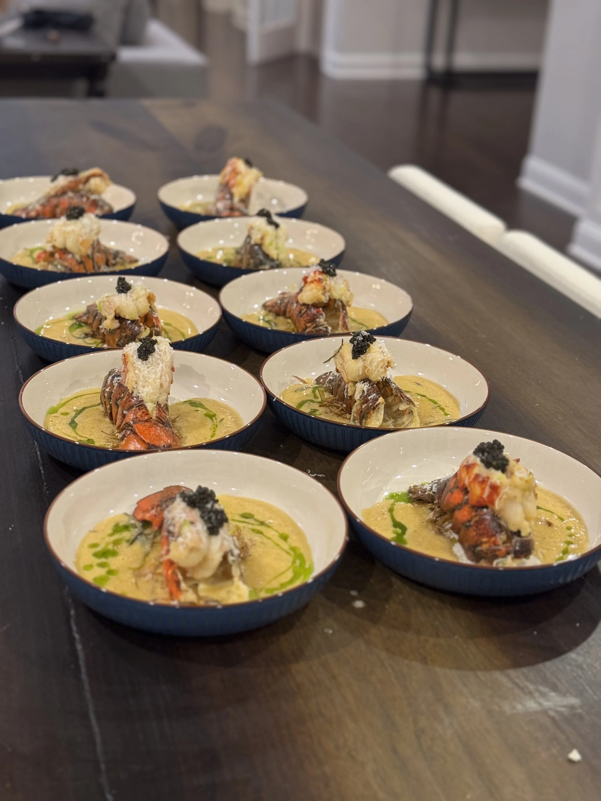Multiple small bowls with lobster tail and sauce, garnished with herbs and caviar, placed on a wooden table.