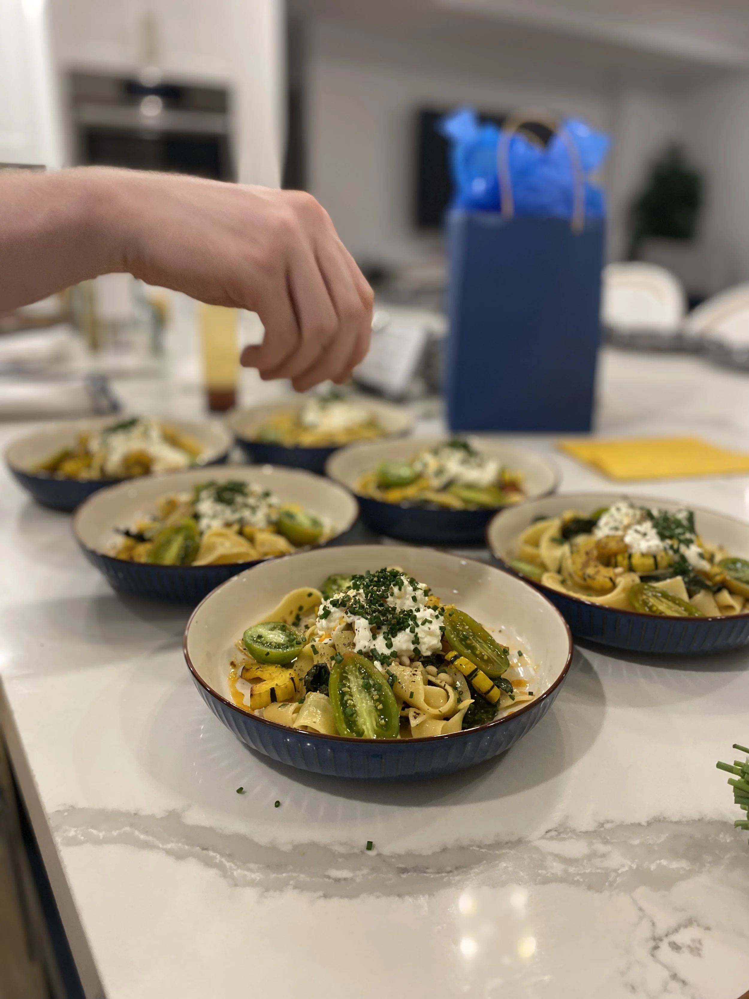 A person preparing bowls of pasta with vegetables and garnishes in a kitchen.
