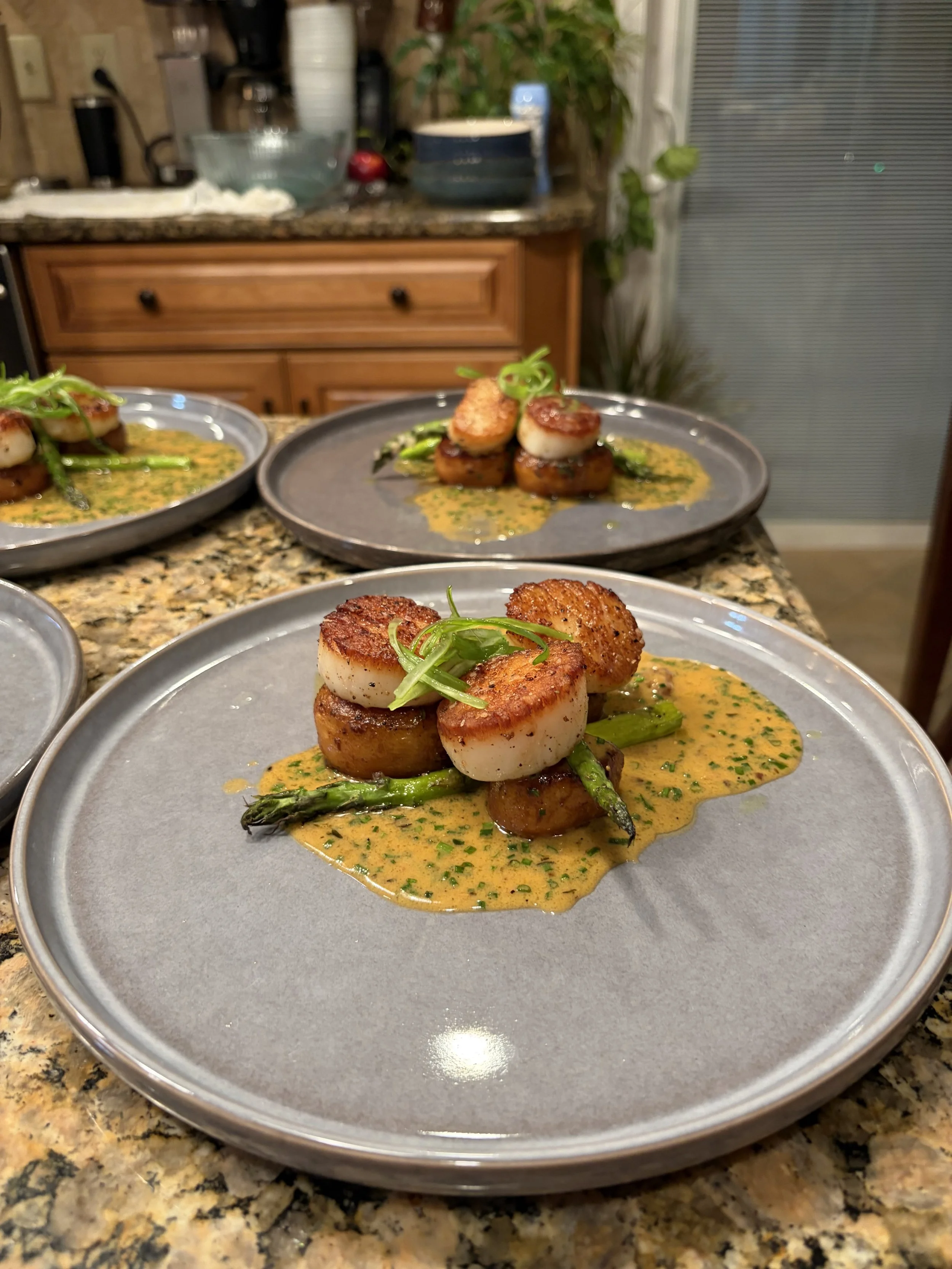 Close-up of a plated dish with seared scallops, grilled asparagus, and a vibrant yellow sauce garnished with microgreens, set on a gray plate in a home kitchen.
