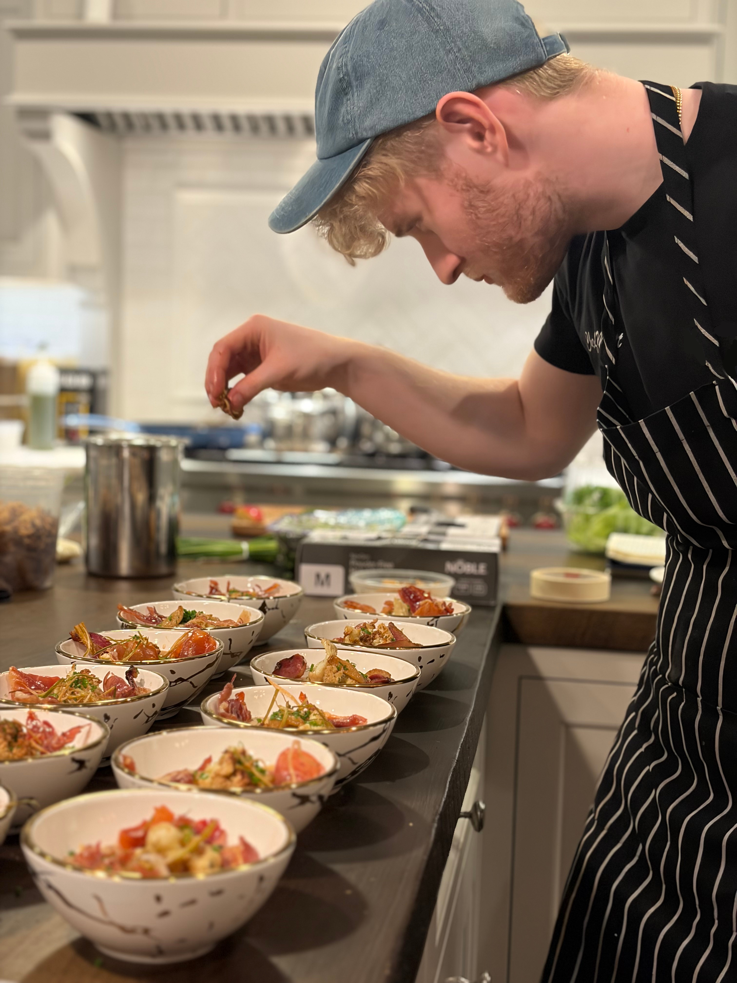 A person wearing a gray hat and striped apron preparing bowls of food on a kitchen counter, with various ingredients and dishes around.
