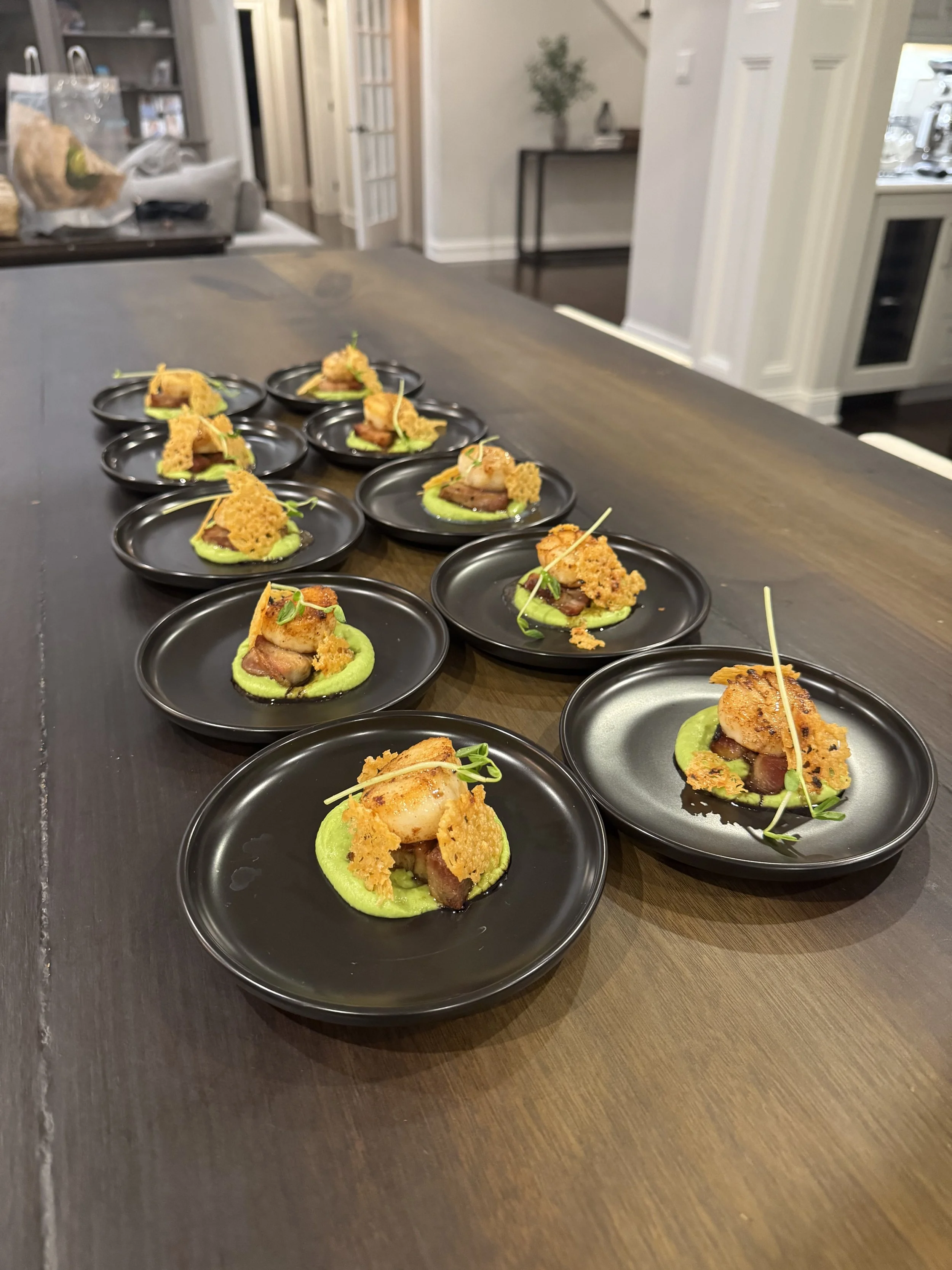 Multiple small black plates each containing a prepared appetizer with a green base, a piece of seared meat, crispy toppings, and garnishes, arranged on a dark wooden table inside a modern kitchen.