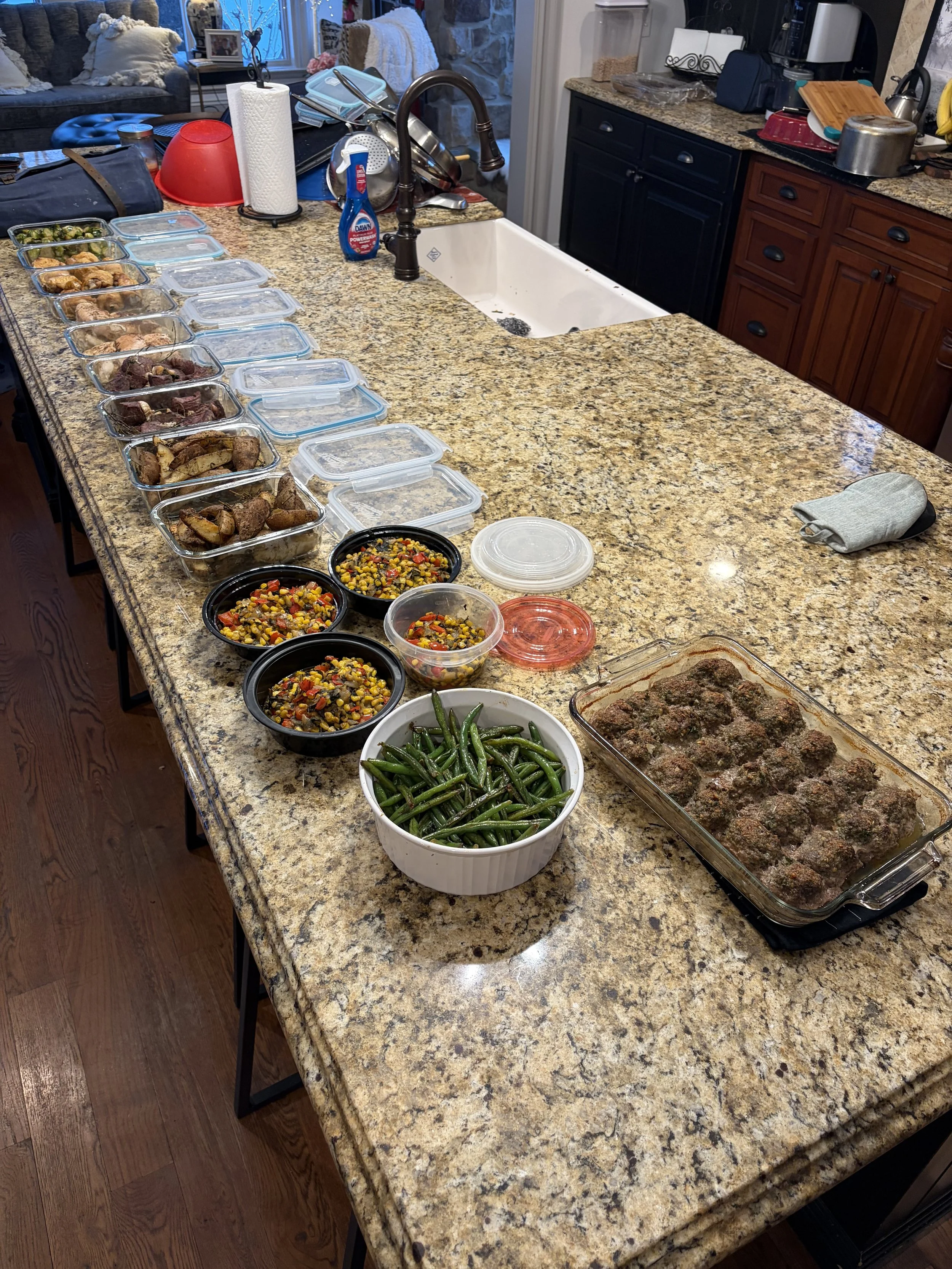 A kitchen island with multiple containers of prepared food, including green beans, meatballs, and mixed vegetables, along with some kitchen utensils and appliances visible on the countertops.