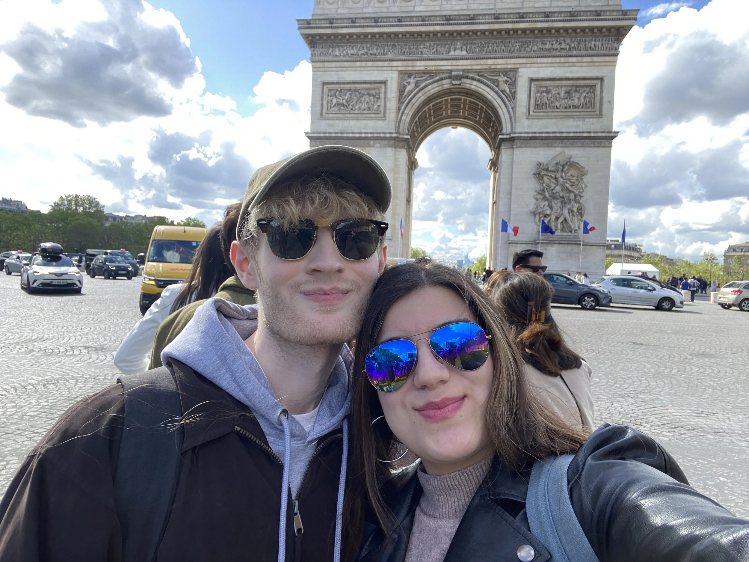 People taking a selfie in front of the Arc de Triomphe in Paris, France, on a partly cloudy day.