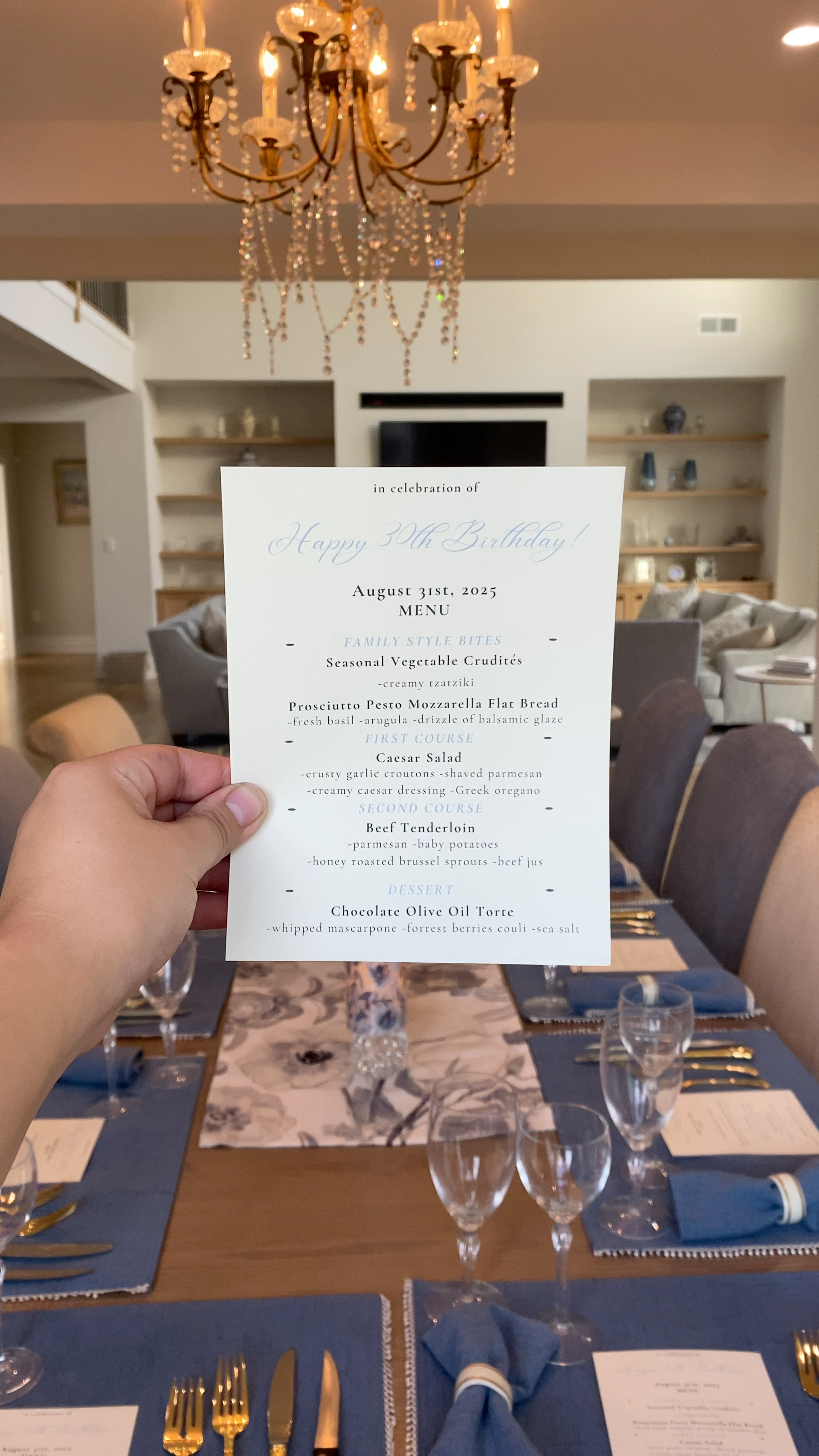 A hand holding a birthday dinner menu for a celebration in a dining room with a chandelier and a dining table set with glasses, gold utensils, and blue napkins.