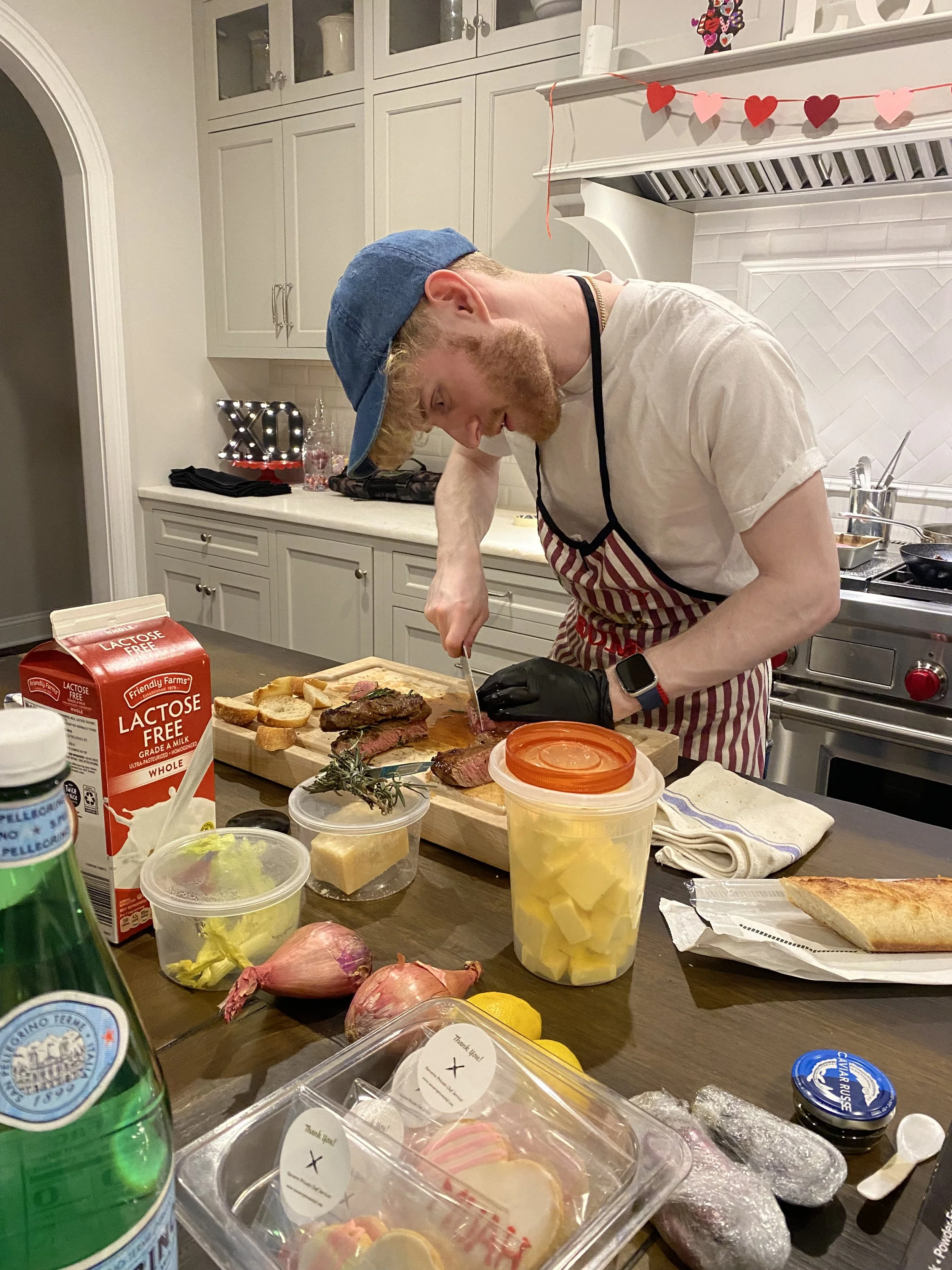 A man with a beard wearing a blue cap, a white t-shirt, an apron, and a smartwatch is slicing a cooked steak on a wooden cutting board in a kitchen decorated for Valentine's Day with heart-shaped garlands. Various food items, containers, and drinks a