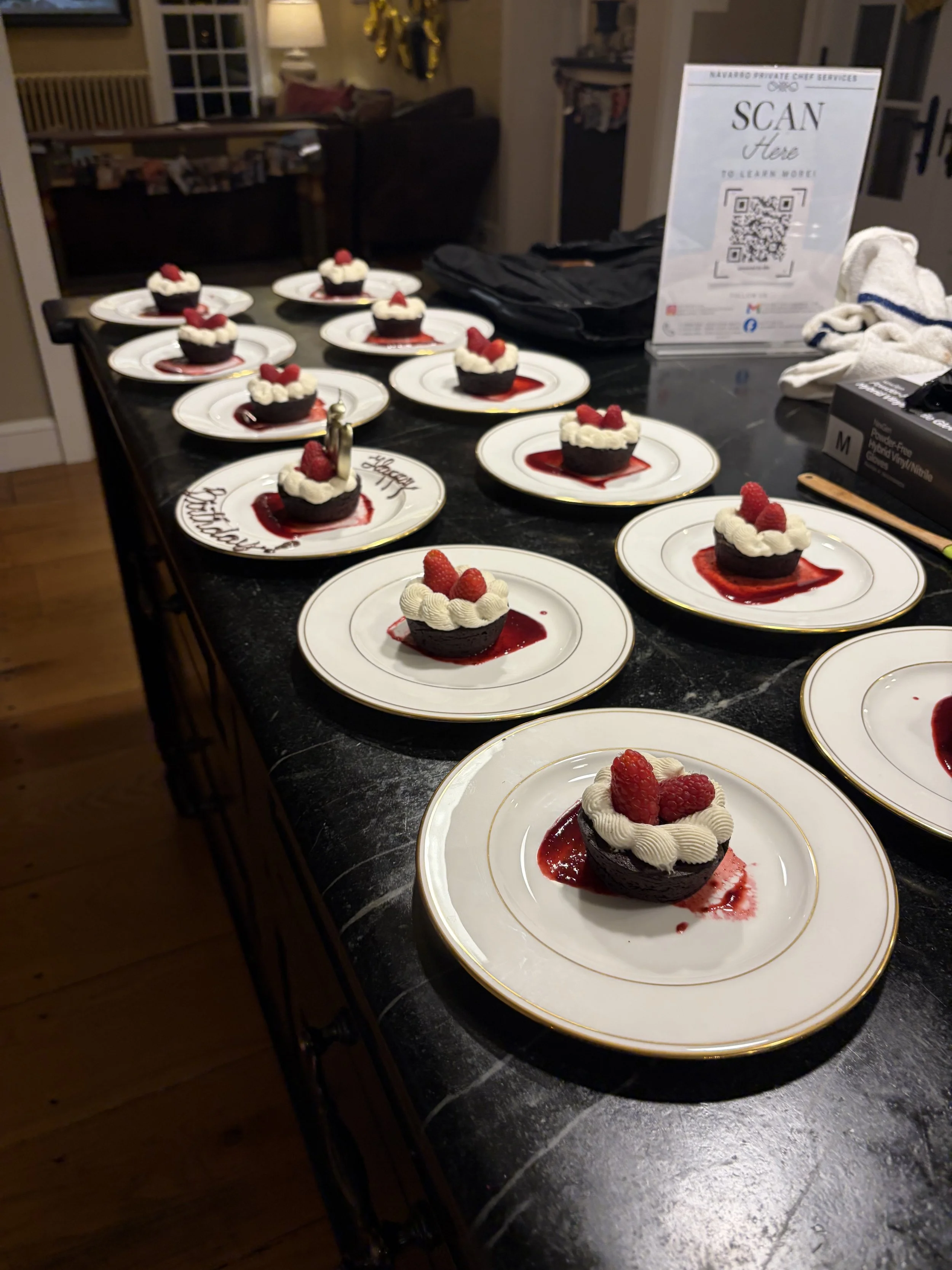 A tabletop with several plates of individual chocolate desserts topped with whipped cream and raspberries, arranged for a celebration in a home setting.