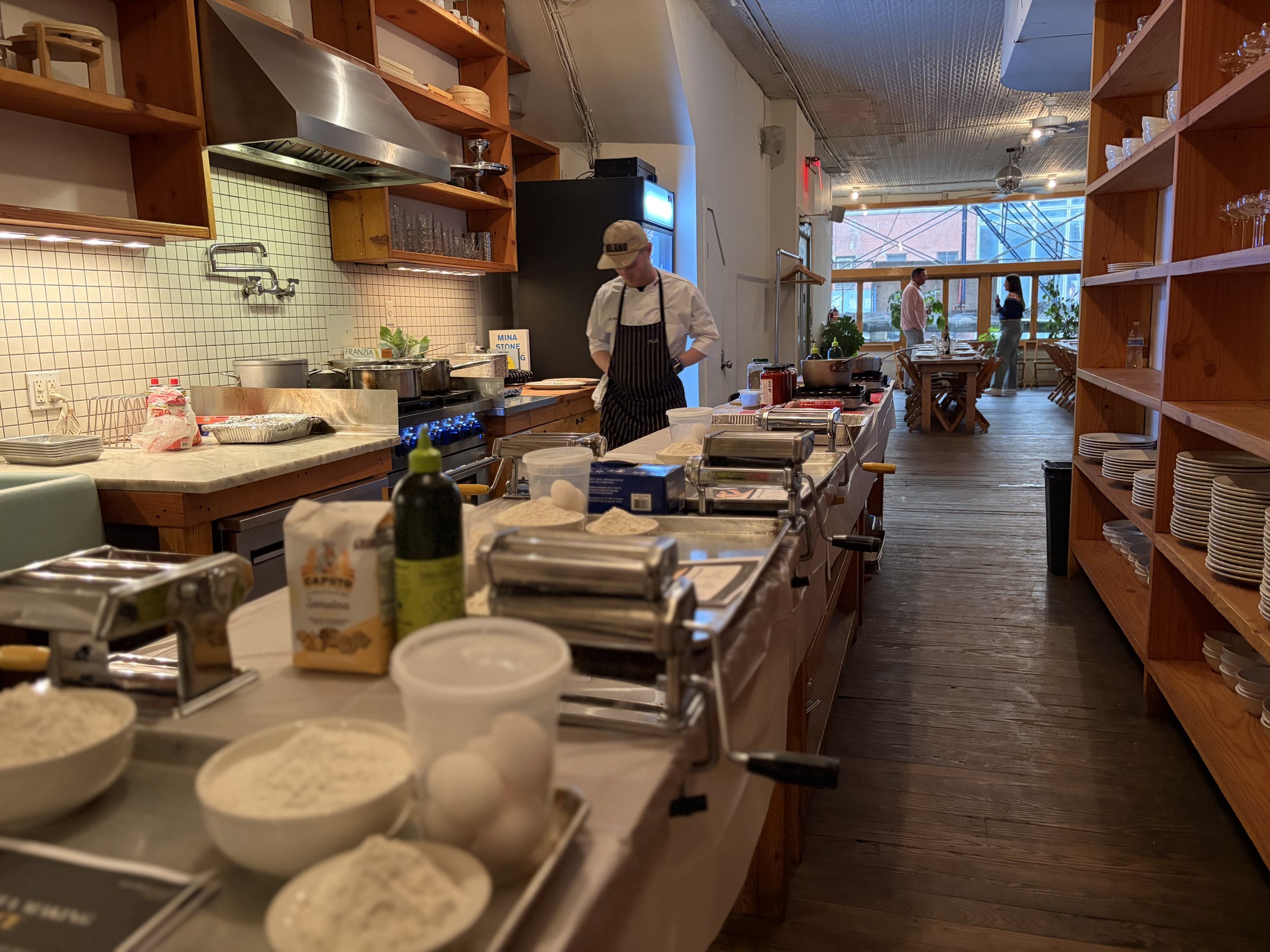 A chef preparing food in a modern kitchen with wooden shelves, large windows, and a dining area with two people talking.