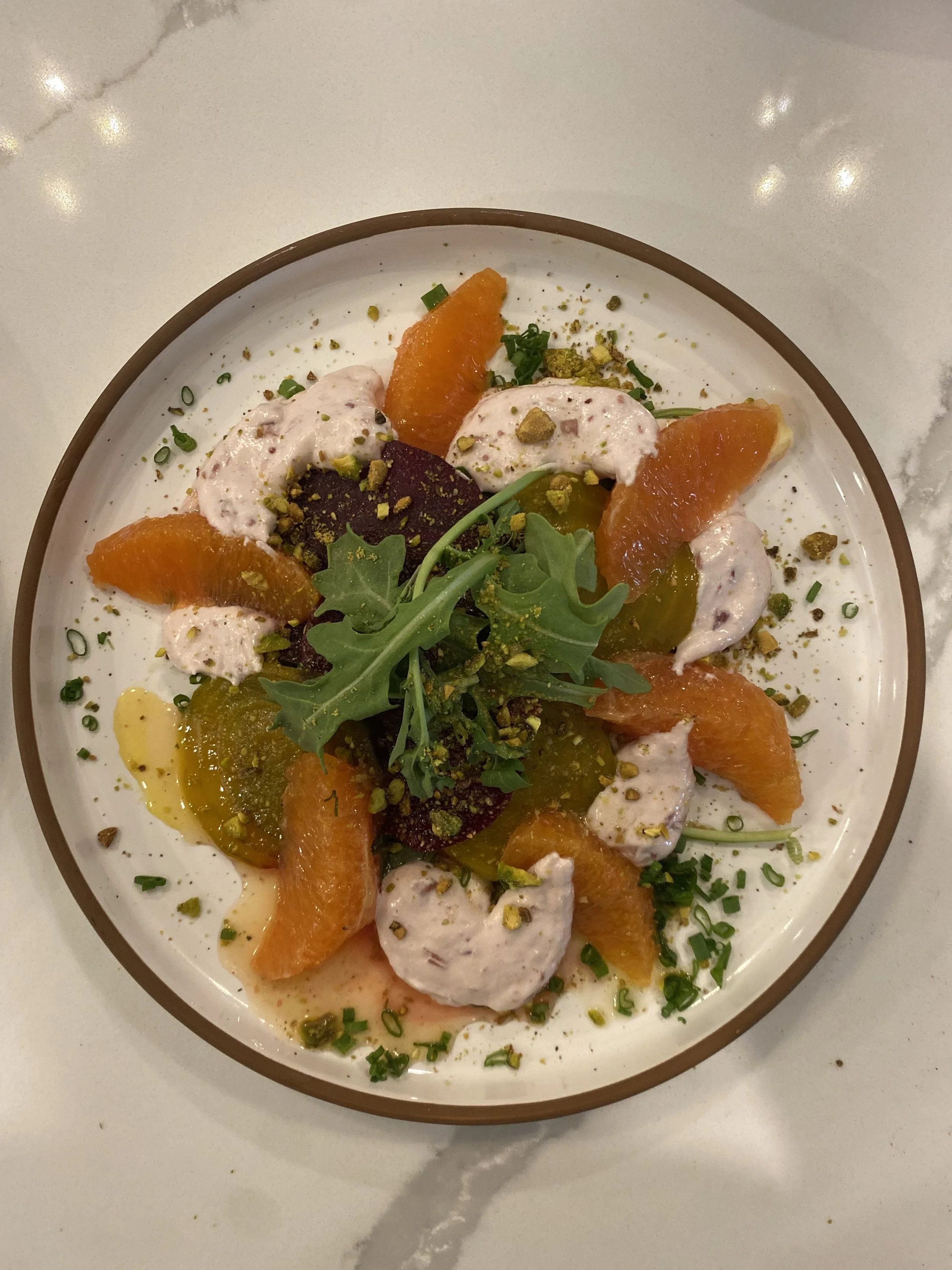A bowl of creamy dessert topped with orange segments, chopped pistachios, and chopped green onions, garnished with arugula leaves.