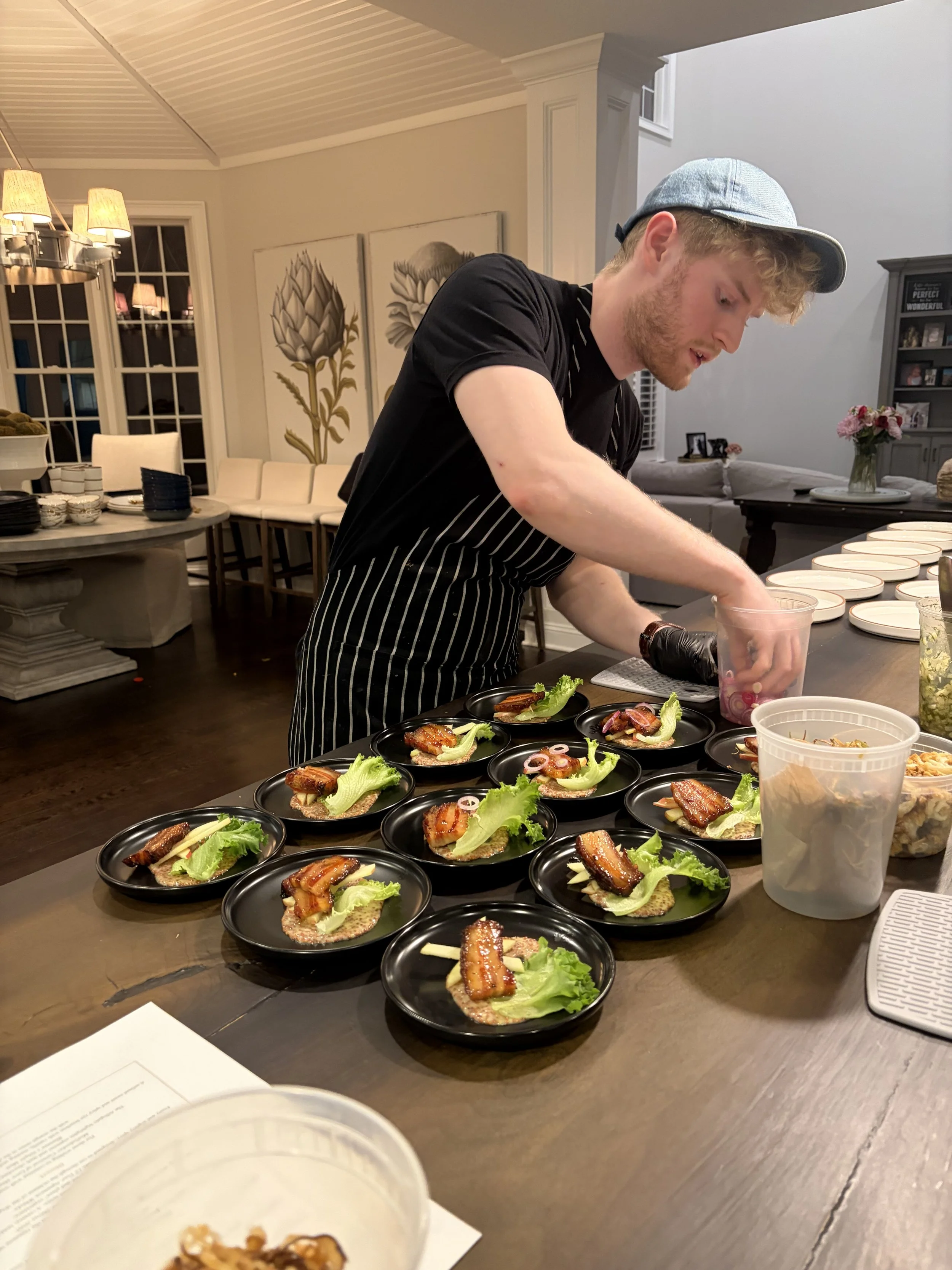 A man preparing small plates of food on a dark wooden table in a cozy, well-decorated living room. The plates contain lettuce, slices of meat, and other toppings. There are containers of ingredients and a vase of pink flowers in the background.