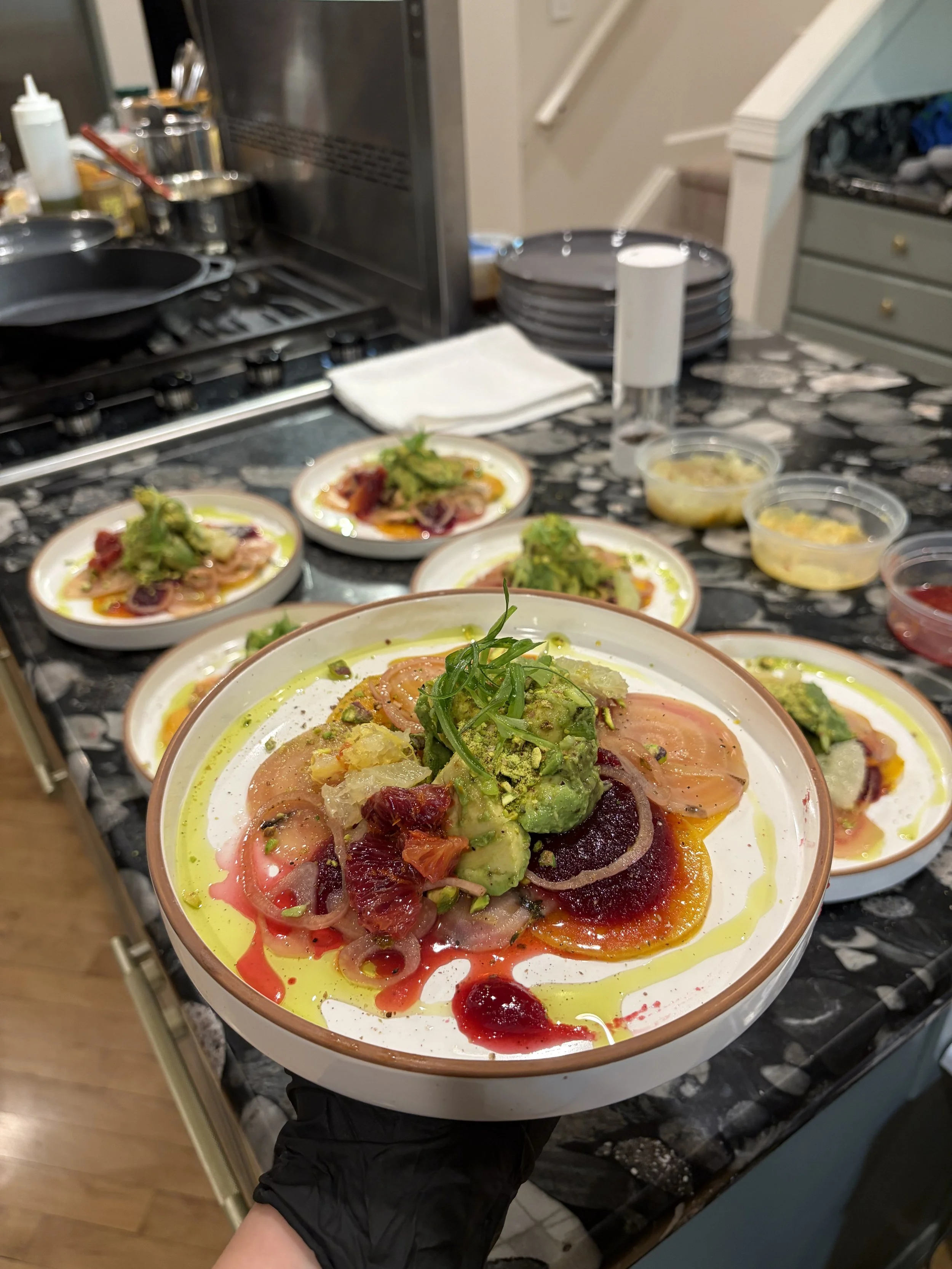 Close-up of a person holding a bowl of colorful, intricately arranged sashimi with slices of fish, avocado, and garnishes, in a kitchen setting with additional bowls and plates visible in the background.
