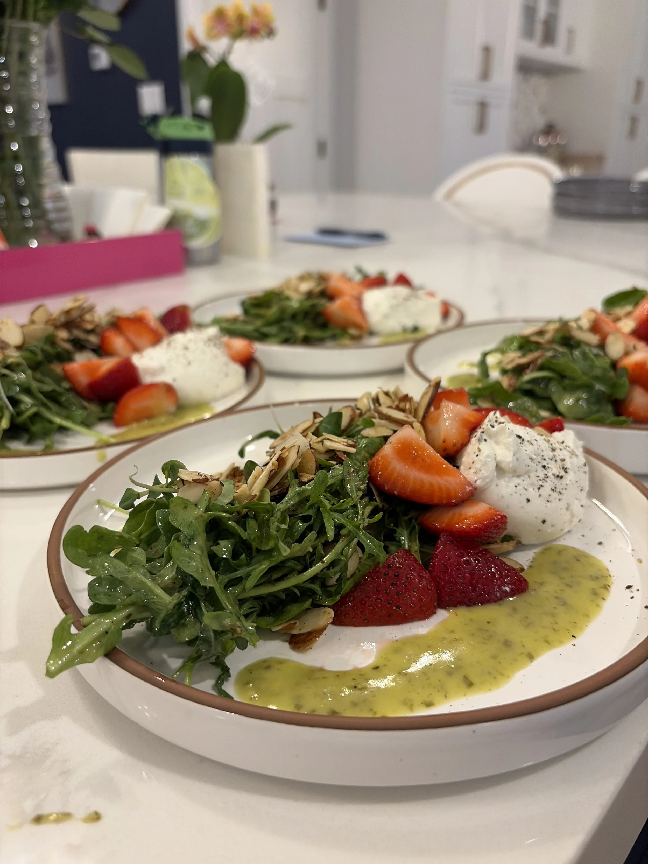A close-up of a plate with a fresh salad including arugula, strawberries, sliced almonds, and a dollop of cream cheese or burrata cheese, drizzled with green pesto sauce. Other plates with similar salads can be seen in the background on a white table