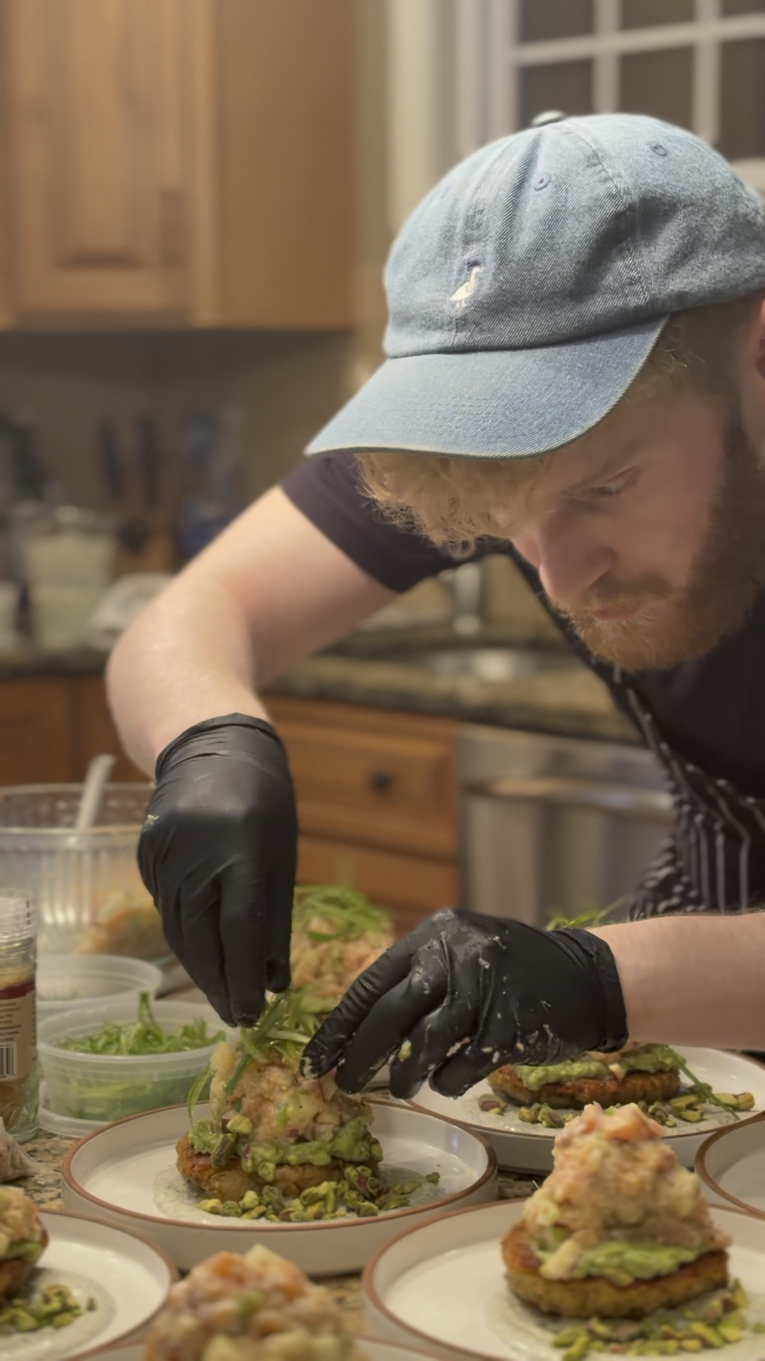 A man in a gray cap and black gloves preparing dishes with green vegetables in a kitchen.