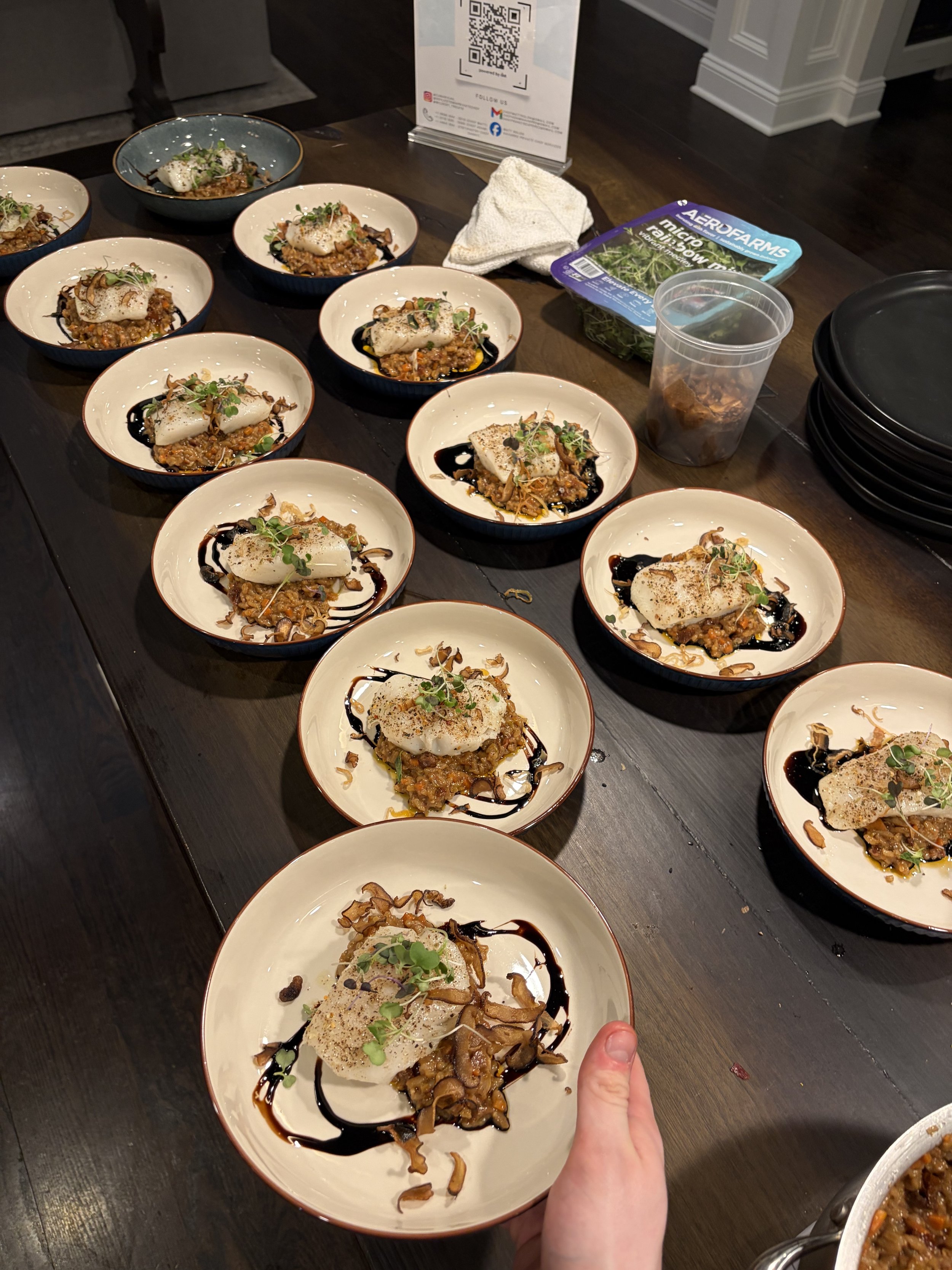 A person holding a plate of food with a thumb showing in the foreground, multiple identical plates of a gourmet dish with microgreens, sauce drizzles, and garnishes on a dark wooden table.