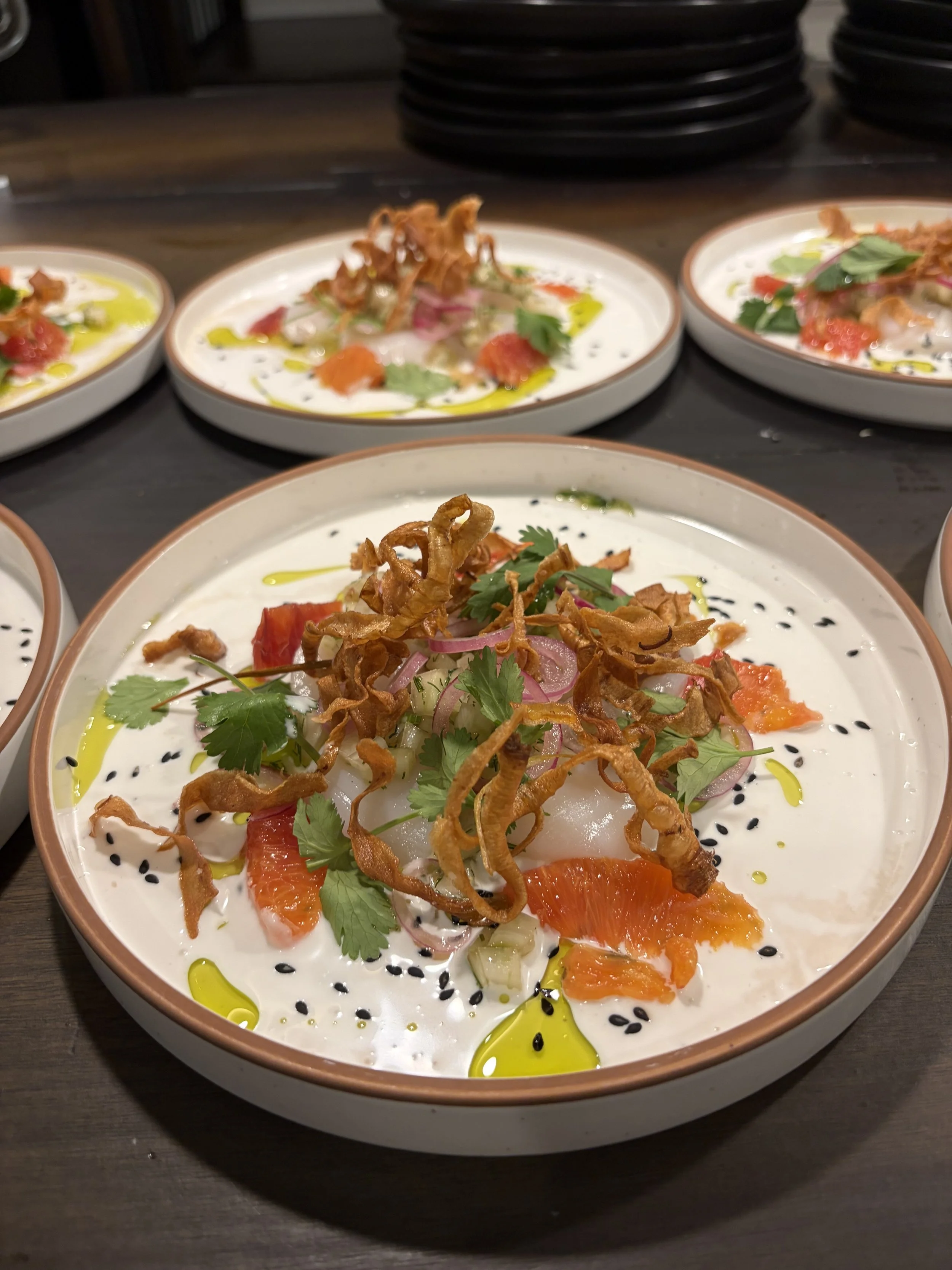 A plate of ceviche garnished with fried shallots, cilantro, sliced red onions, and orange segments, drizzled with green and black sesame seeds and olive oil.