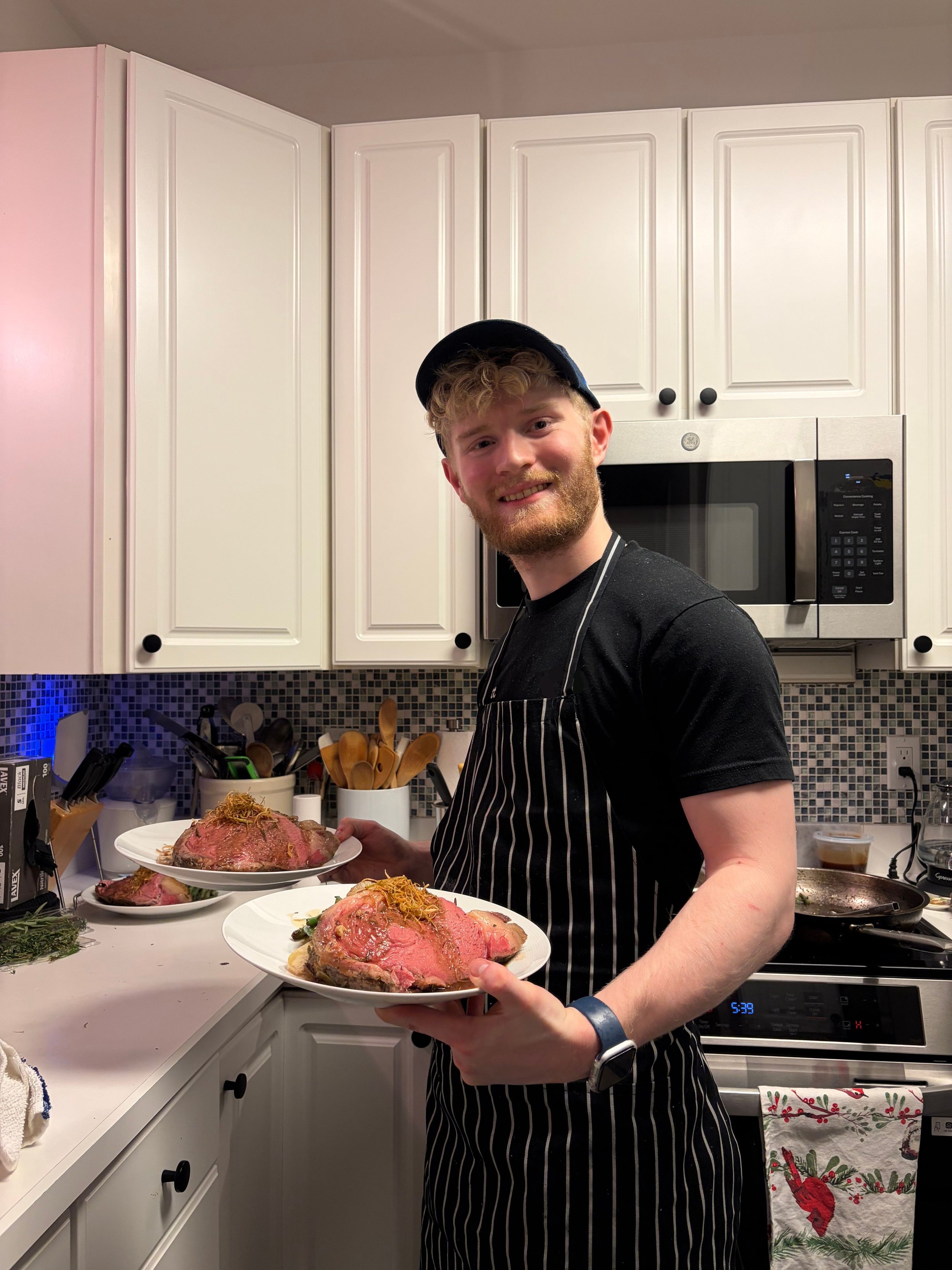 A young man with curly blond hair and a beard, wearing a black cap and a black striped apron, is smiling and holding a plate with a large roast beef. The kitchen has white cabinets, an oven, and various cooking utensils on the counter.