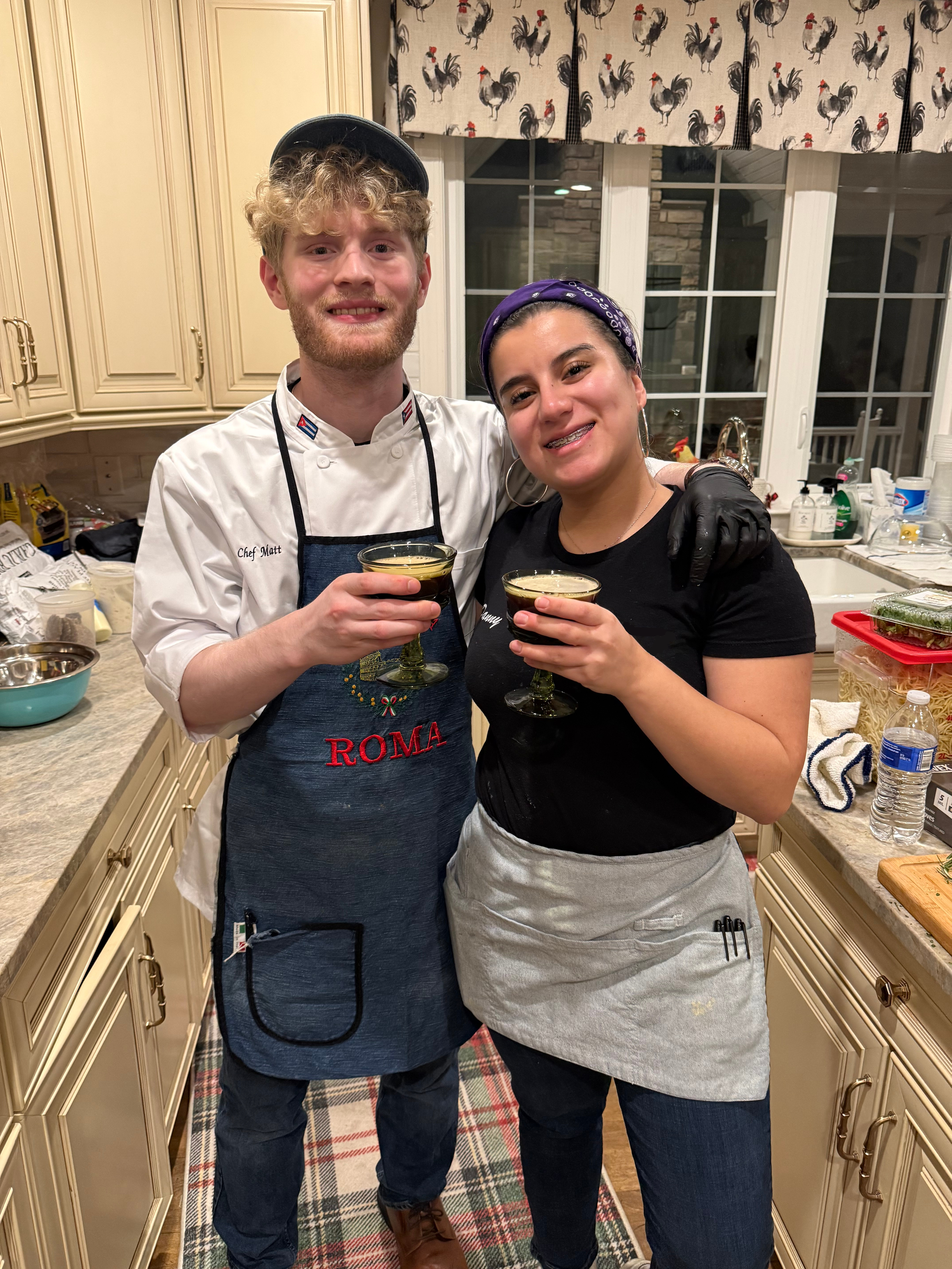 Two people in a kitchen, smiling and holding glasses of drinks. One person is dressed as a chef with a white coat and apron, and the other is dressed casually with a headscarf and hoop earrings. The kitchen has cream-colored cabinets, a window with roosters on the curtains, and various kitchen items on the countertops.