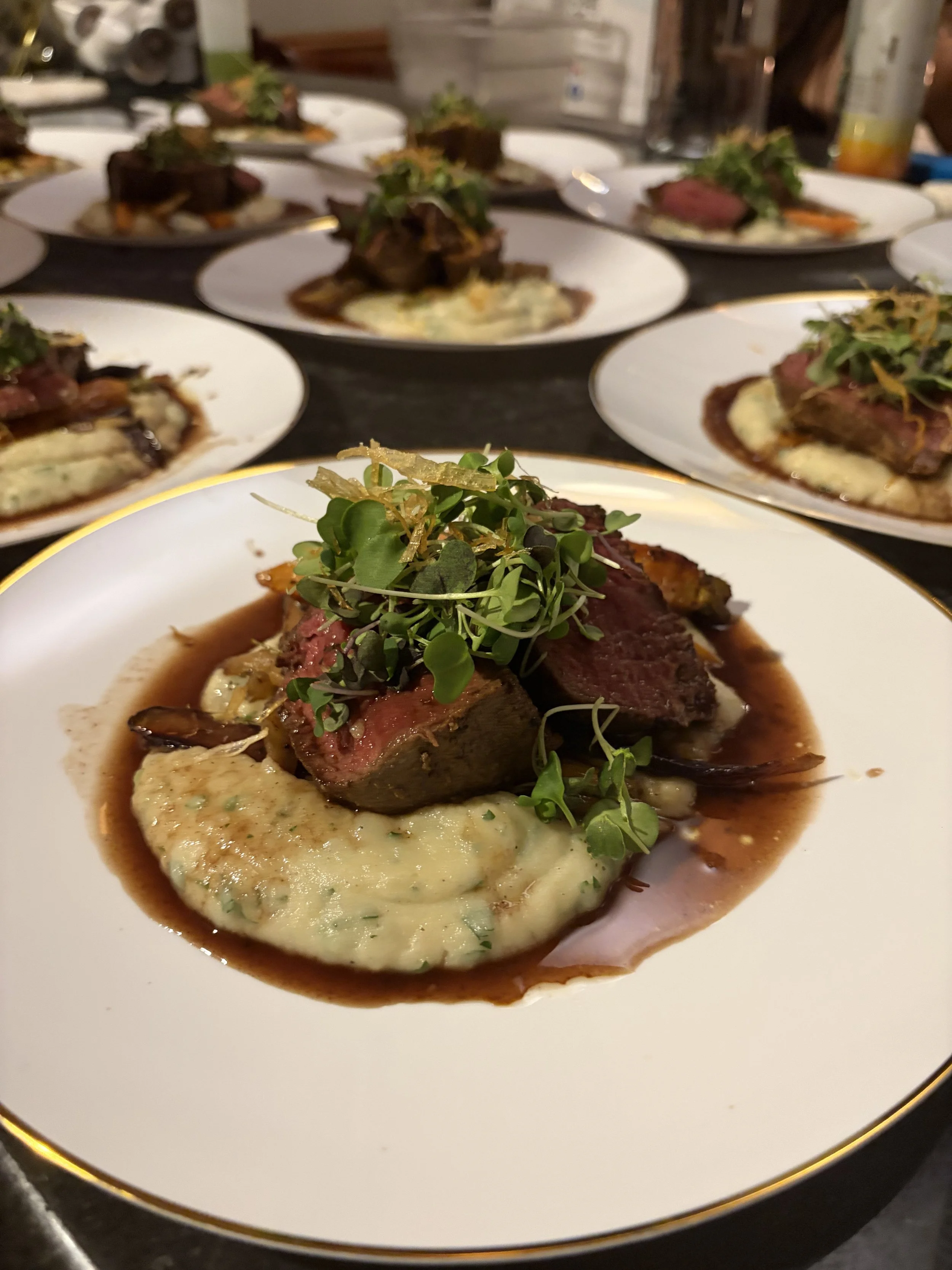 A plated dish of beef steak topped with microgreens, served with mashed potatoes and a brown sauce on a white plate with a gold rim.