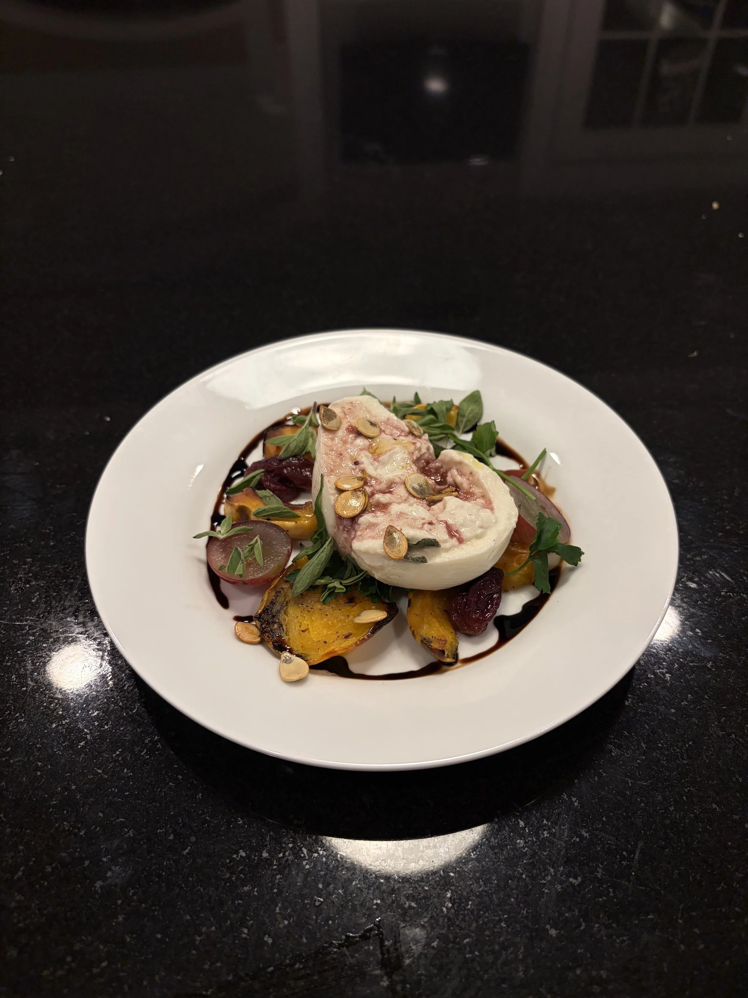 A plated dish with a piece of creamy cheese topped with pumpkin seeds, garnished with fresh herbs, and served with roasted vegetables and drizzled with balsamic glaze.