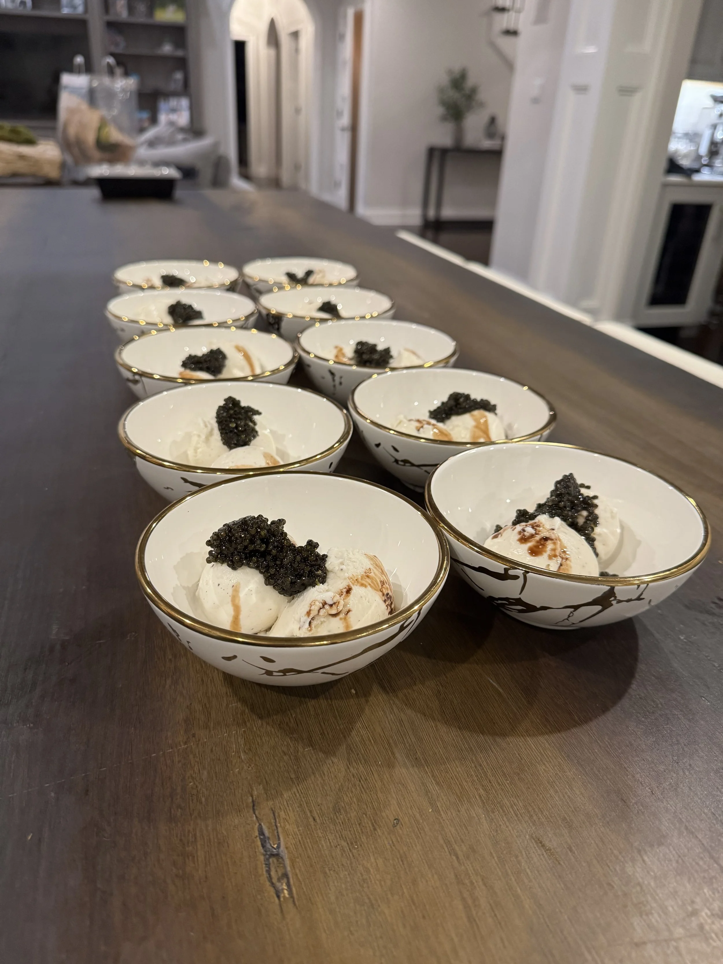 Multiple white porcelain bowls with black marble-like toppings on desserts, arranged on a wooden table in a kitchen.