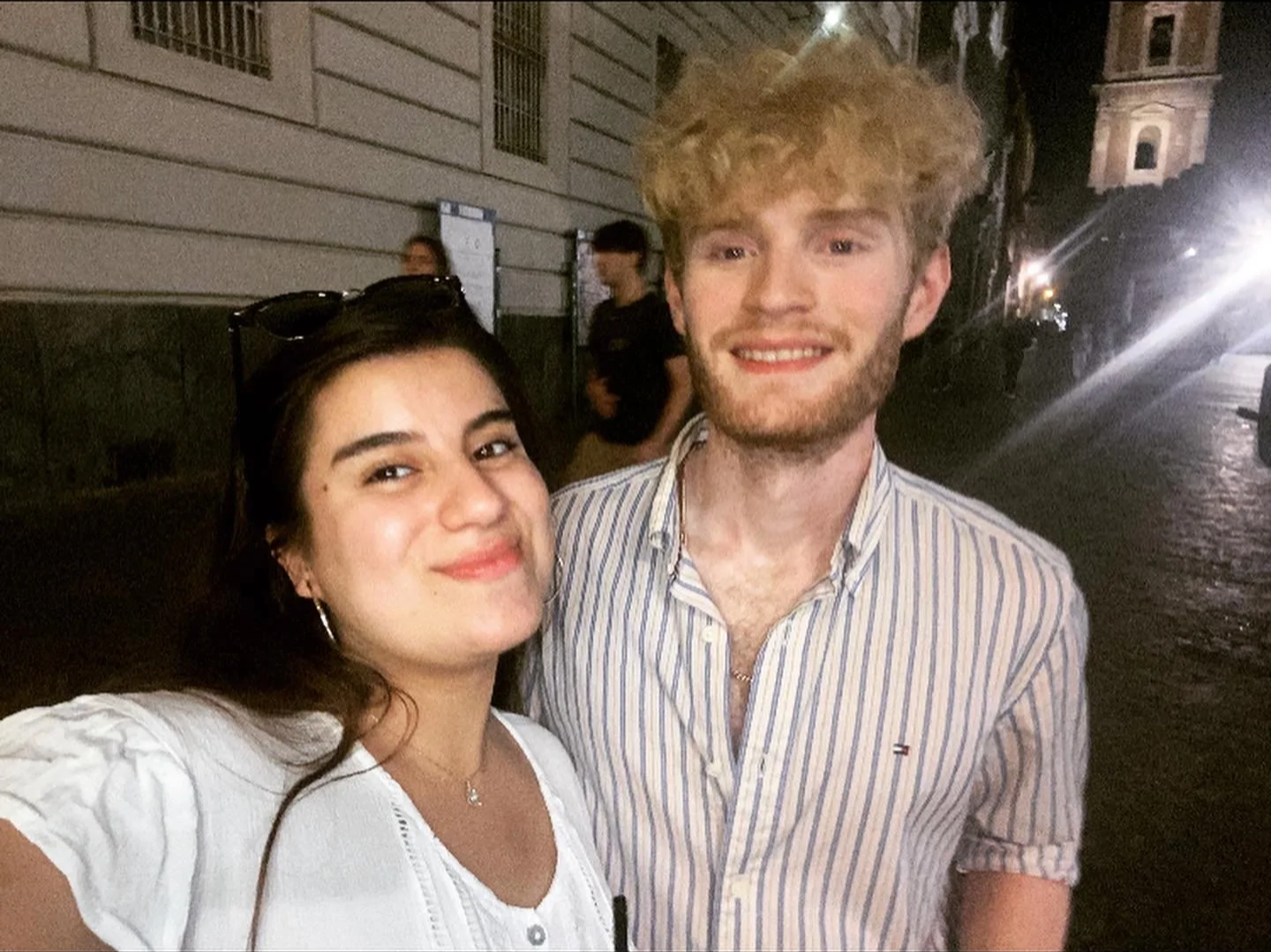 A young woman and a young man taking a selfie on a city street at night, smiling at the camera. The woman has dark hair, tan skin, and is wearing a white top. The man has blonde, curly hair, light skin, and is wearing a striped shirt.