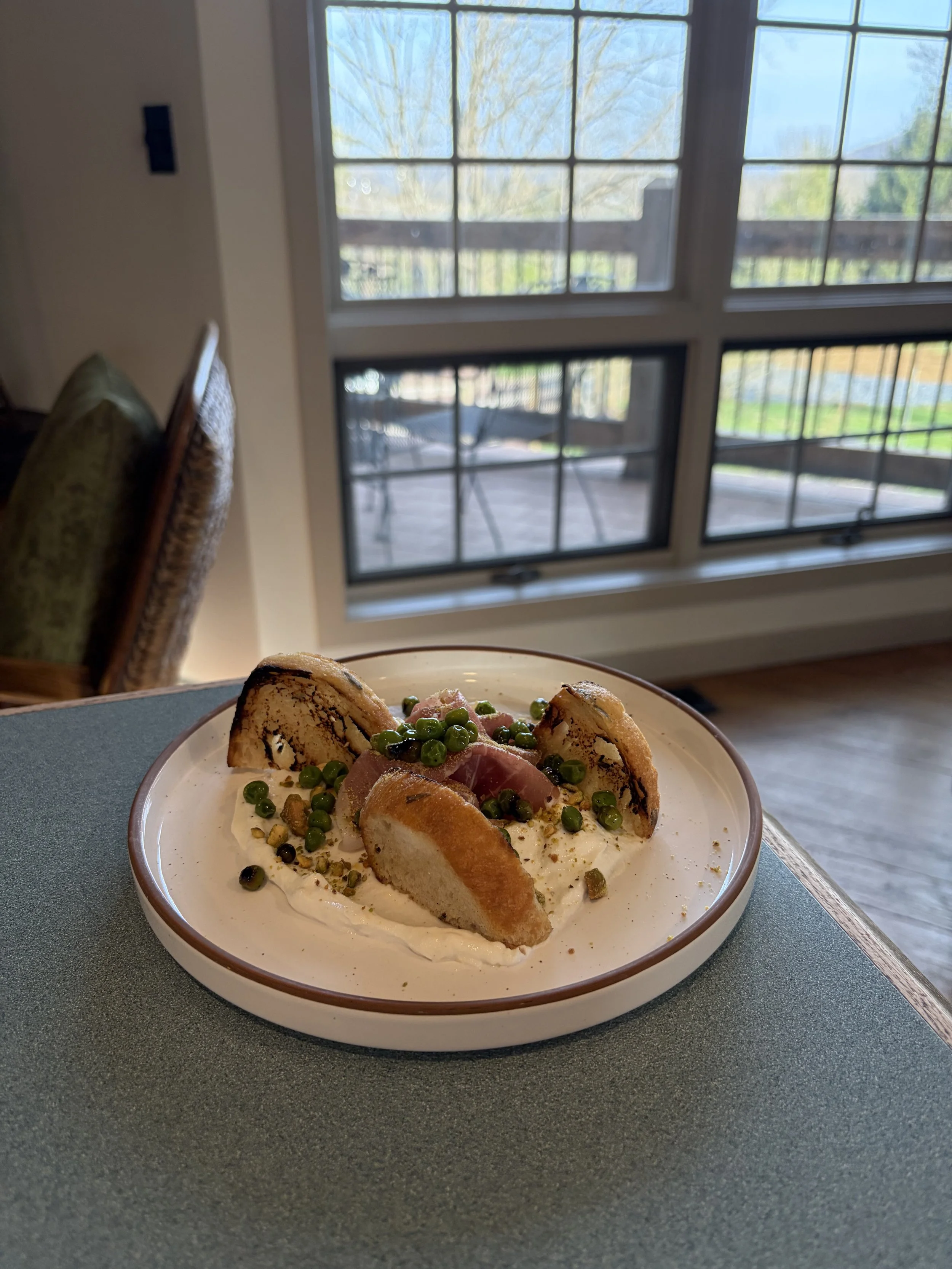 A plate of food with slices of toasted baguette, green peas, and some sliced ham, served with a creamy white sauce, on a table near a window showing an outdoor deck and trees.
