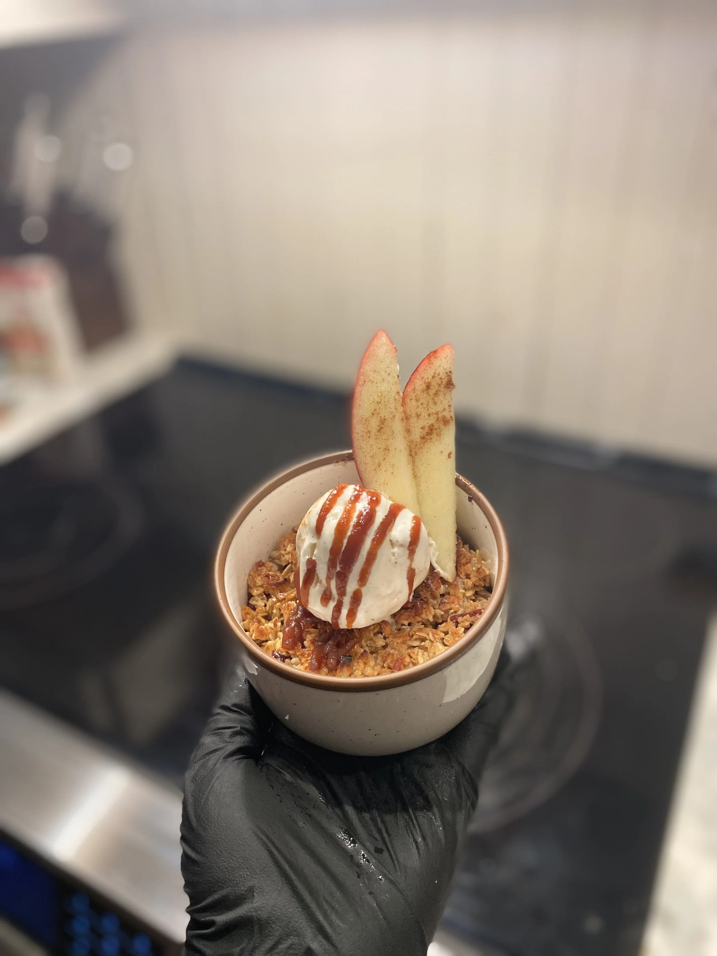 A dessert in a small white bowl with a scoop of vanilla ice cream topped with caramel sauce, garnished with two slices of fresh apple with cinnamon.