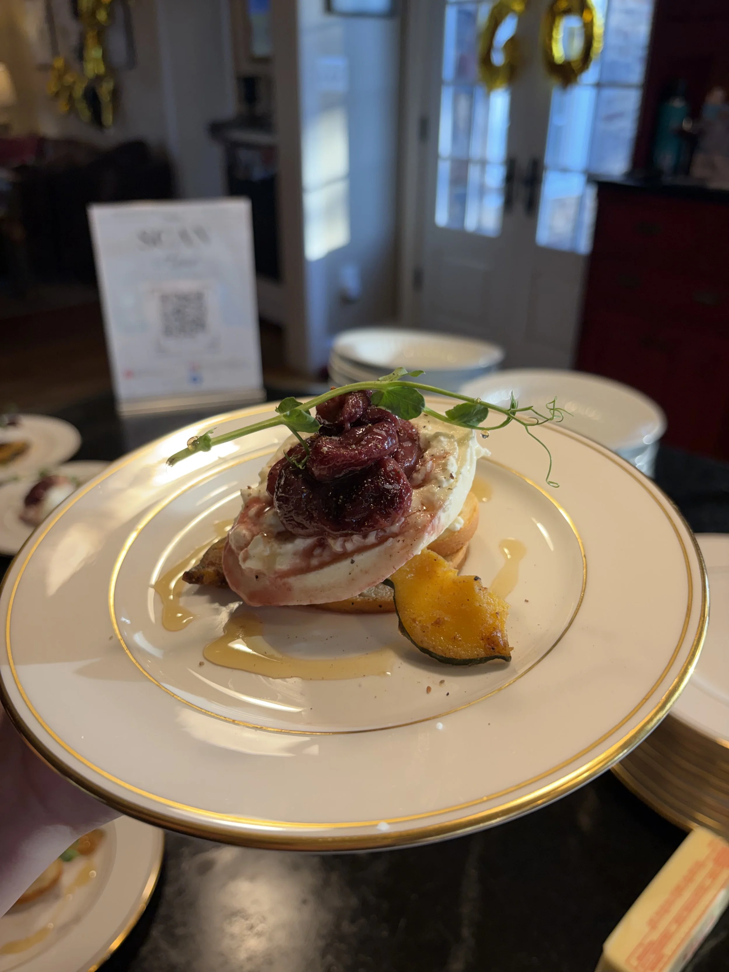 A plate of food with a slice of bread, topped with a dollop of cream, a cherry compote, and a garnish of microgreens, served with a roasted yellow squash wedge, on a white plate with gold trim.