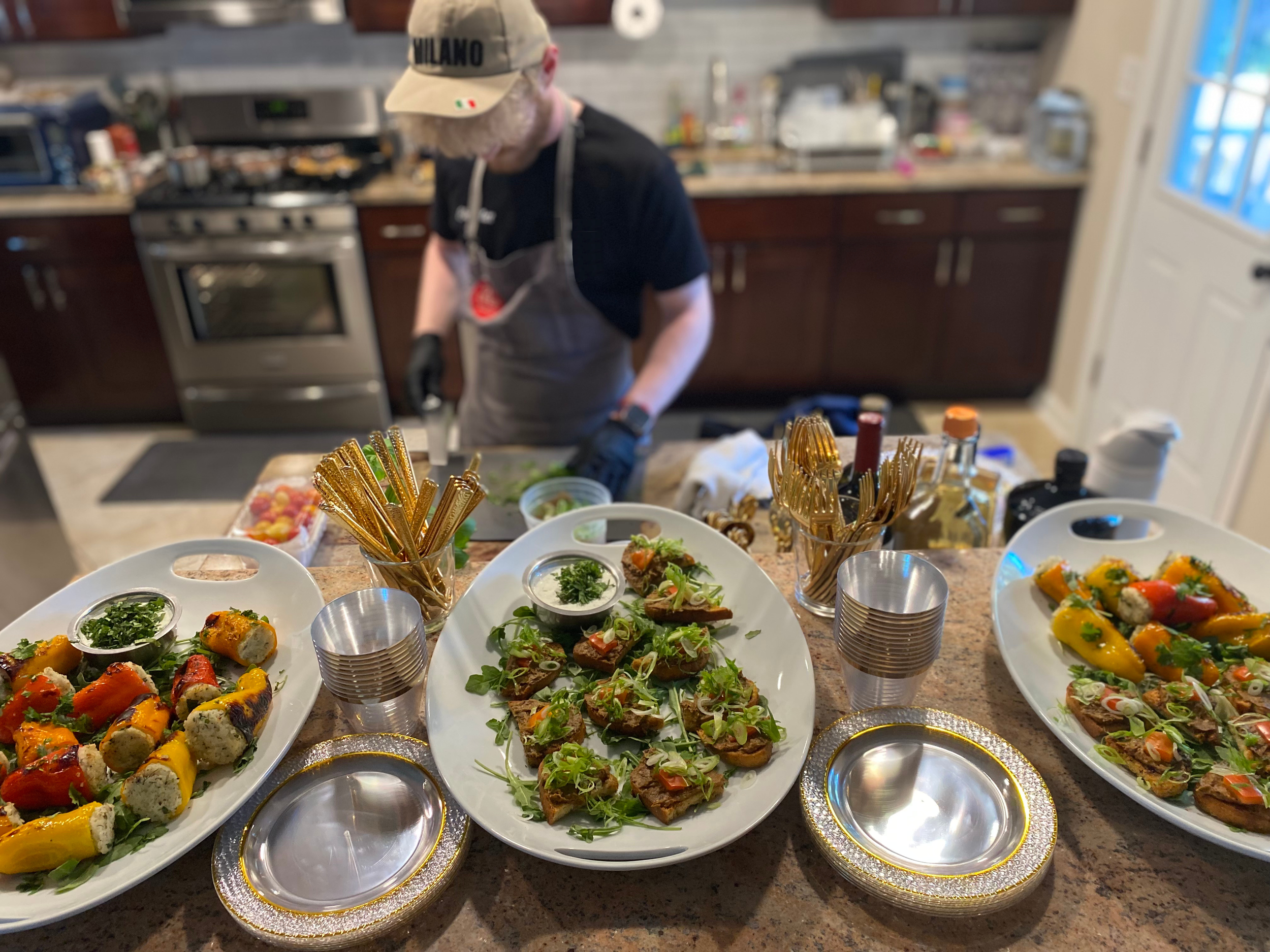 A kitchen with a person preparing food in the background, and a table filled with plates of colorful appetizers, including rolls, sliders, and vegetable dishes, with utensils and condiments in the foreground.