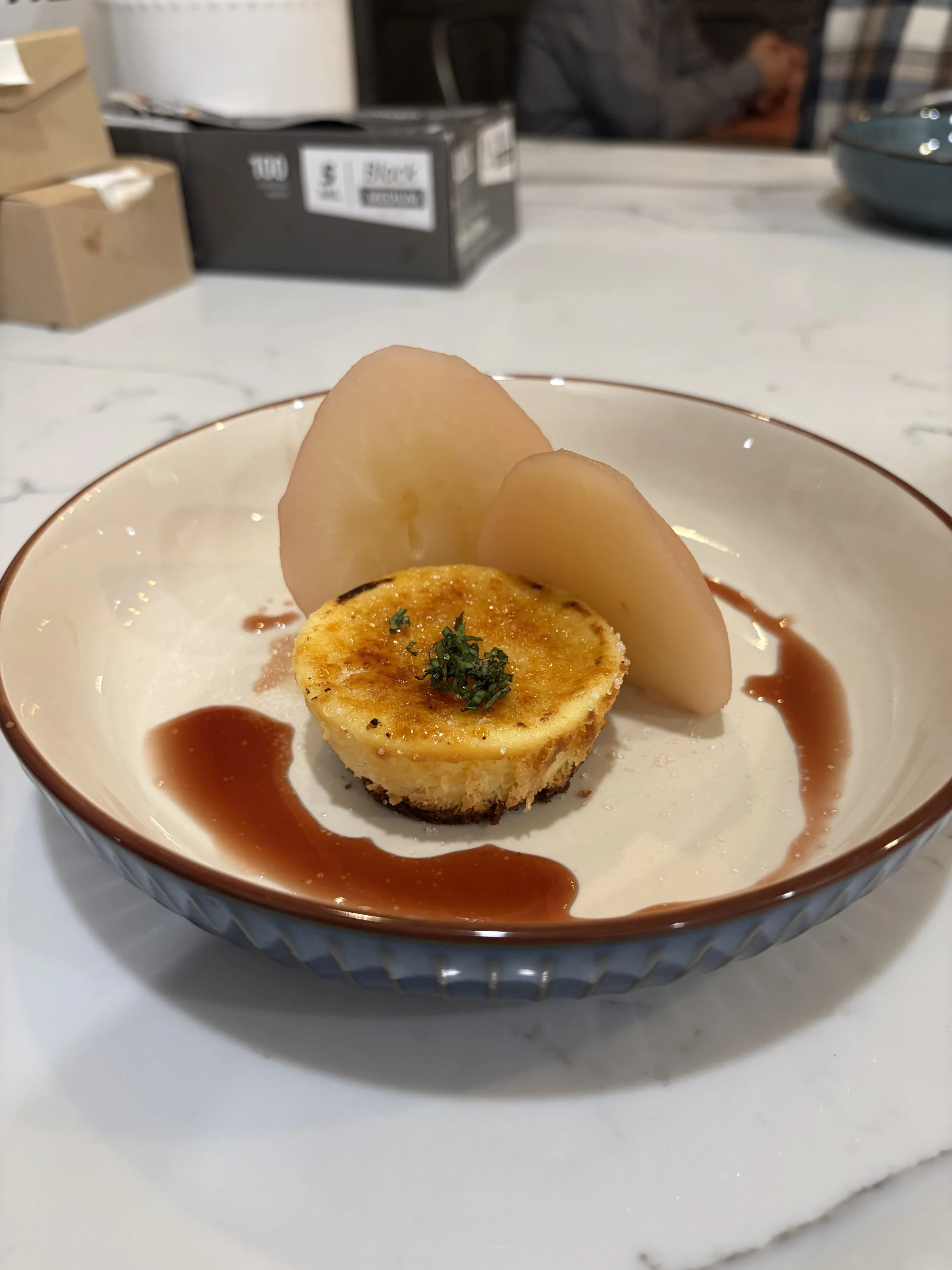 A small round slice of baked potato topped with herbs, served in a white bowl with a gold rim, garnished with two slices of pear and drizzled with sauce on a white marble surface.