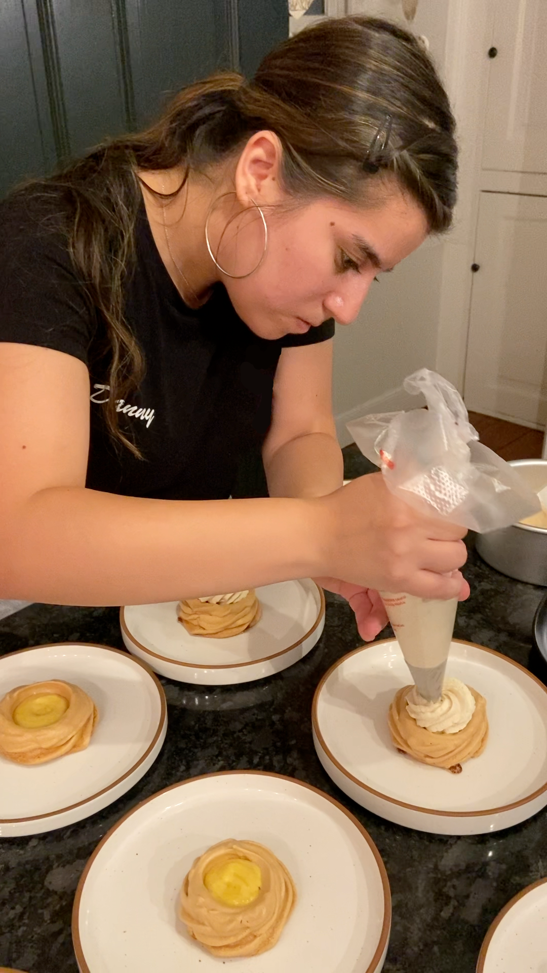 Woman decorating cream-filled pastries with piping bag in a kitchen, surrounded by plates of similar pastries.
