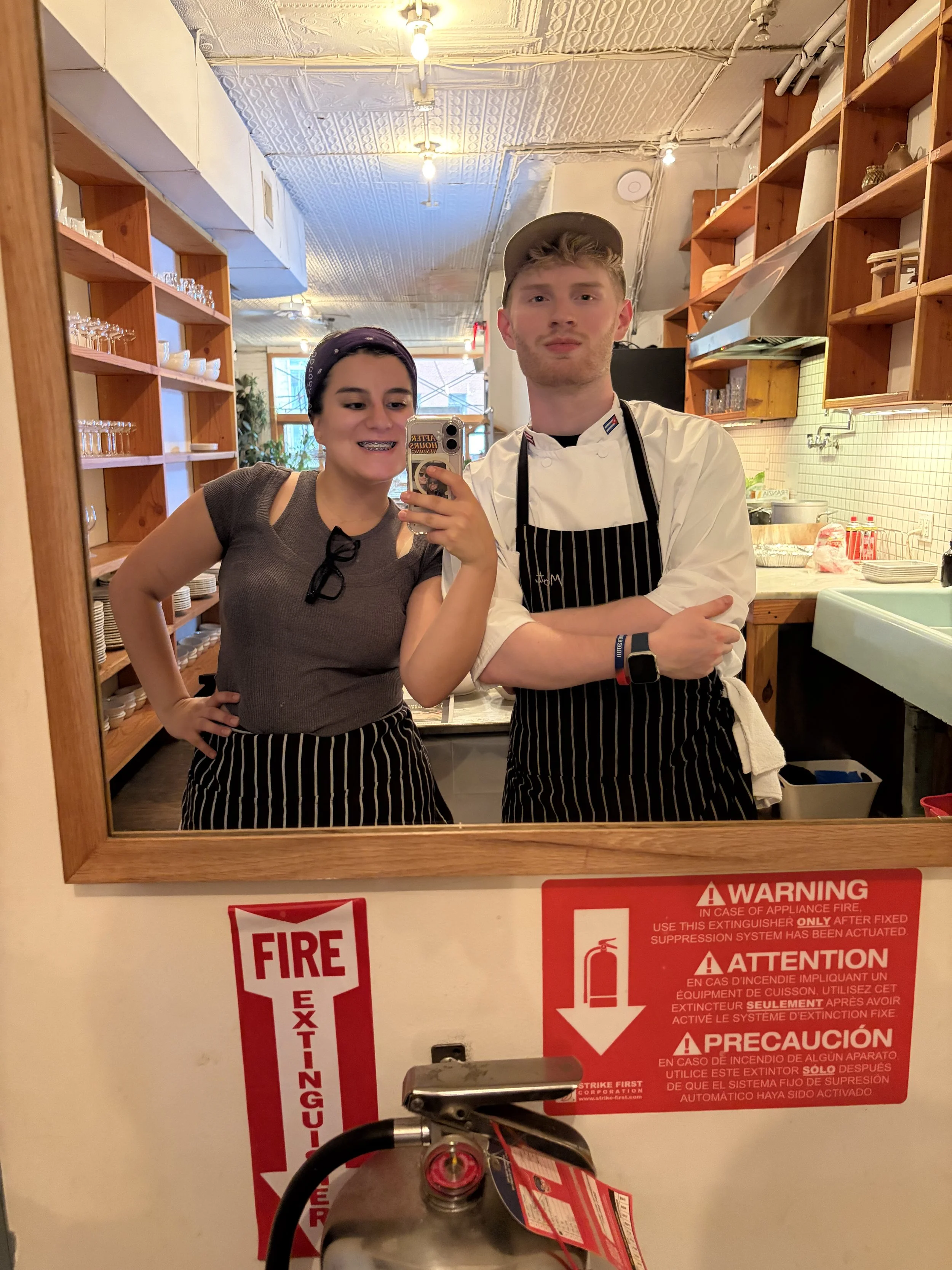 A woman and a man take a selfie in a mirror in a restaurant or cafe. The woman wears a dark top and a purple headscarf; the man is dressed as a chef with a white coat and a striped apron. The background includes shelves with glasses and dishes, and a window with plants outside.