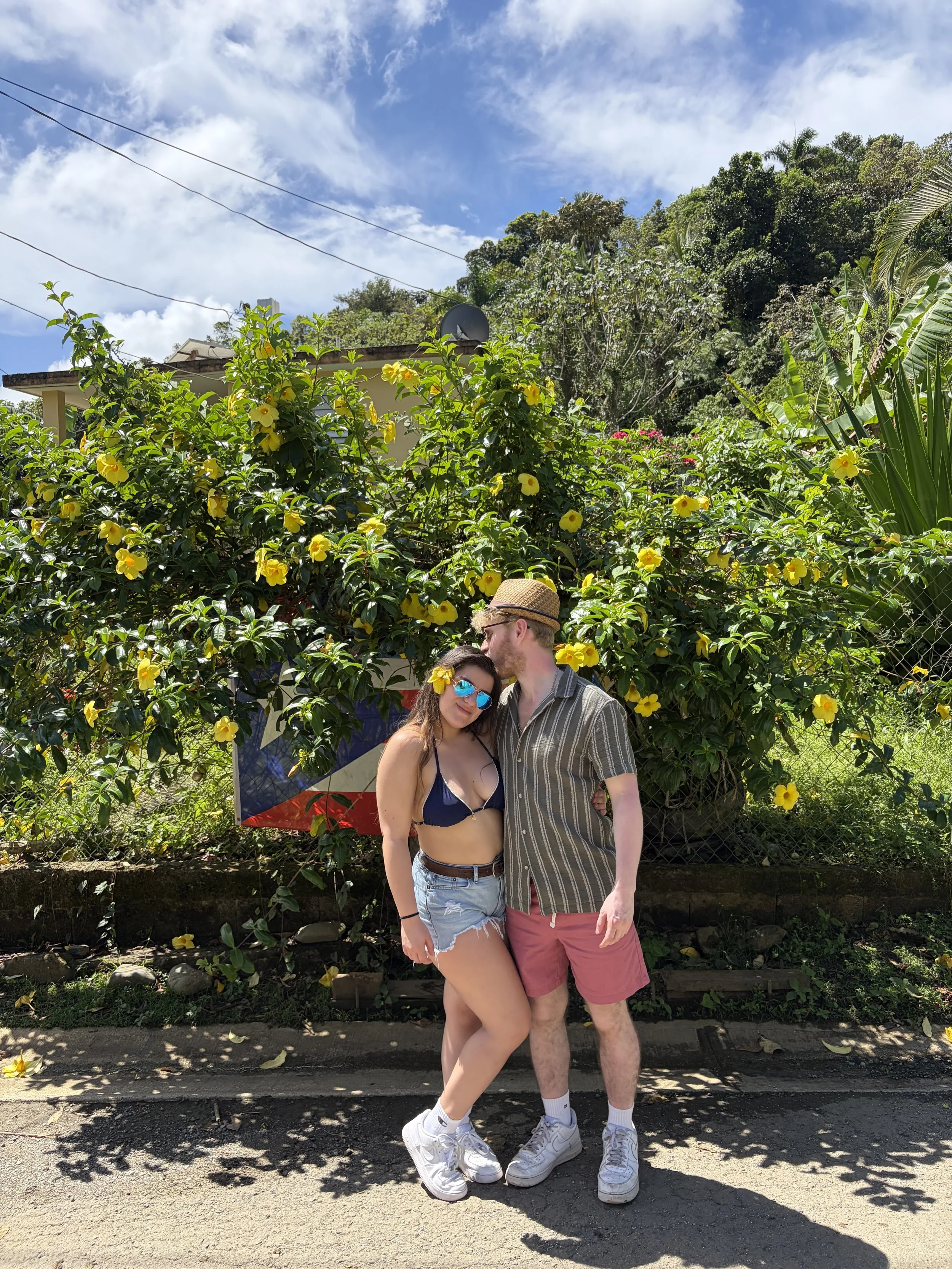 Two people standing together outdoors, a woman wearing sunglasses and a flower in her hair, and a man with a hat, in front of lush greenery and yellow flowers.