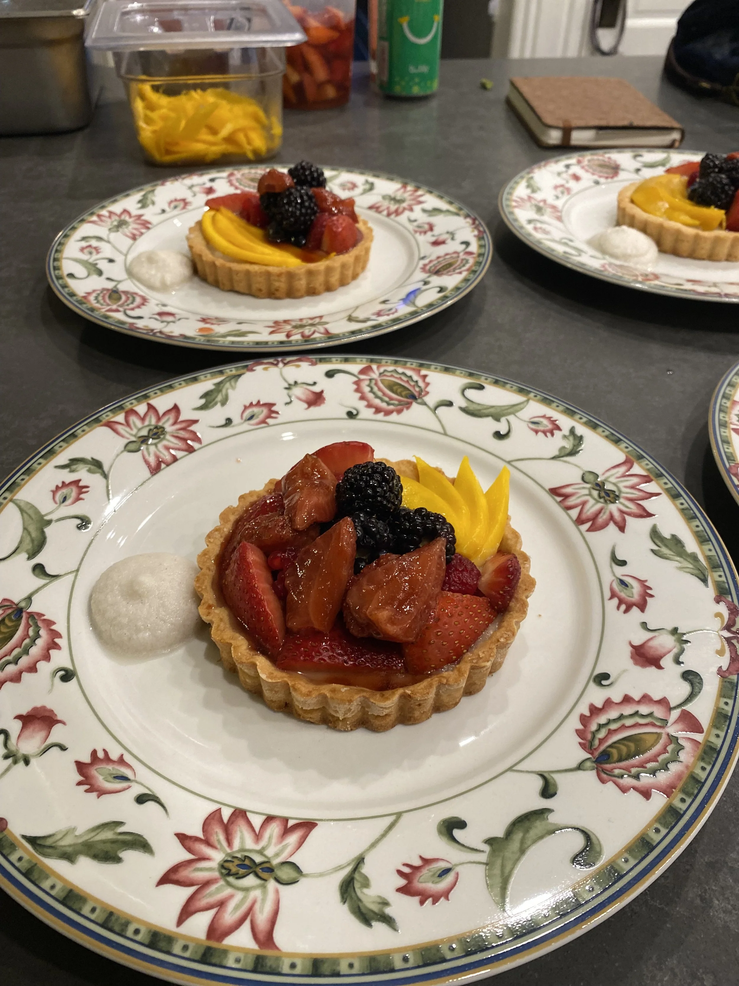 A fruit tart on a decorative floral plate with strawberries, blackberries, and mango slices, accompanied by a dollop of cream.