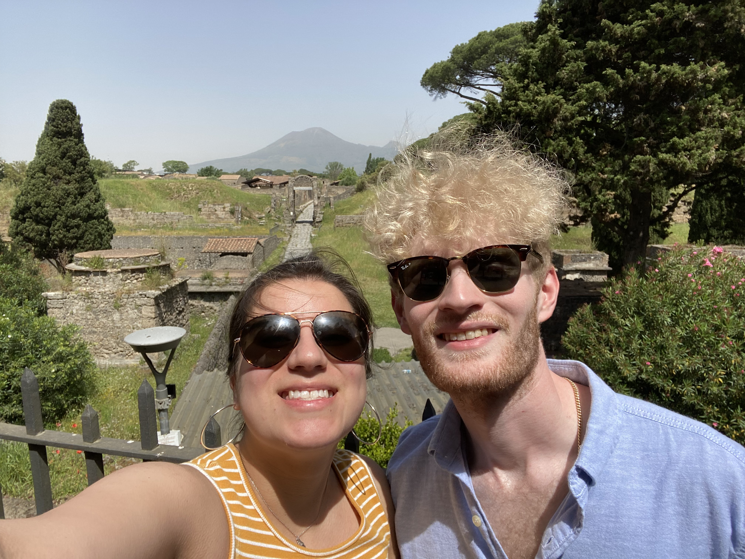 Two people taking a selfie outdoors with mountains and trees in the background. The woman on the left wears sunglasses and a yellow striped top. The man on the right has curly blonde hair, sunglasses, and a light blue shirt.