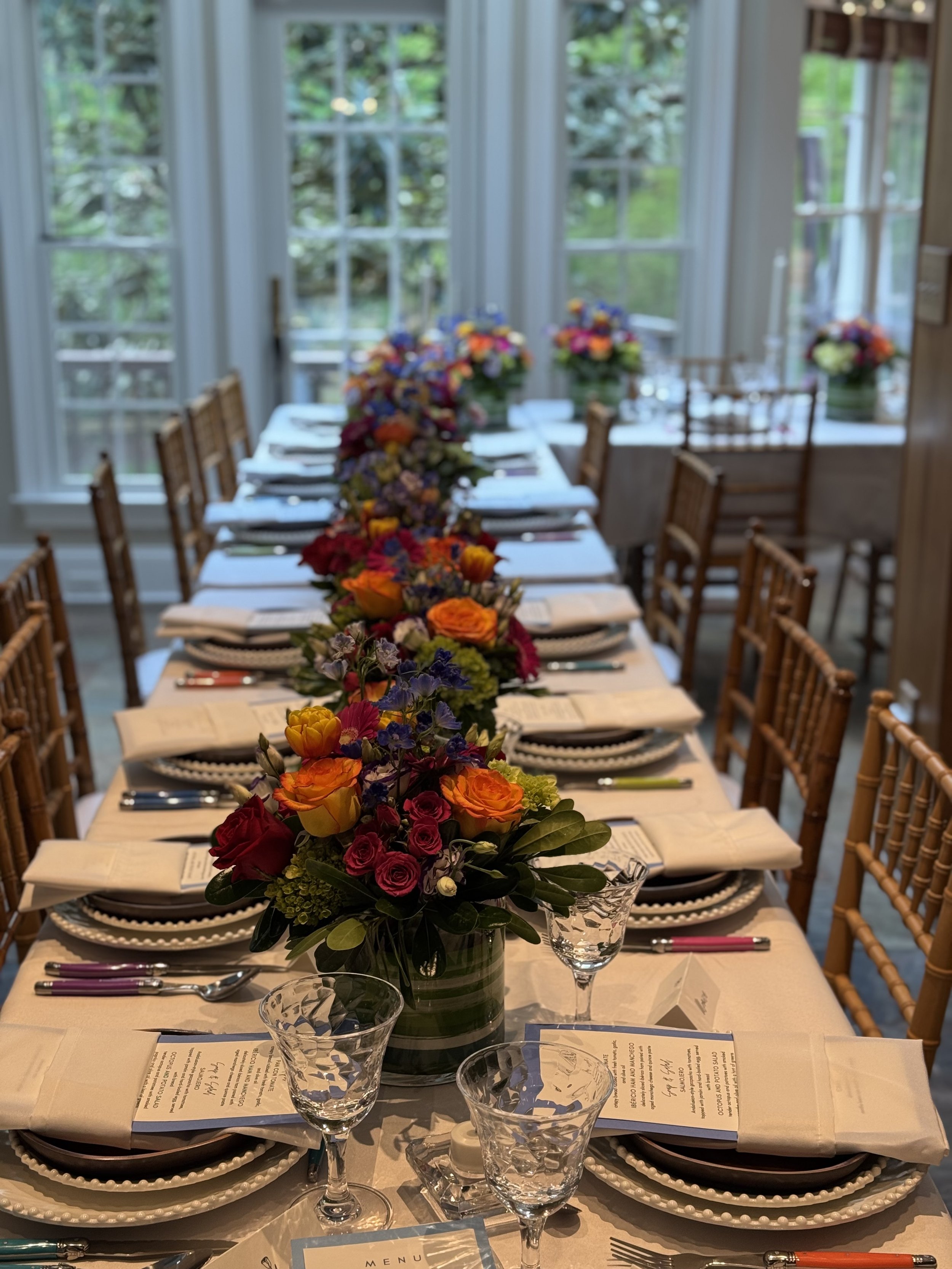 A formal dining table set with plates, forks, knives, purple-handled utensils, menus, empty wine glasses, and glass water glasses. In the center, there is a large bouquet of colorful flowers in a green striped vase with a water-filled glass in front. The background features large windows with greenery outside.