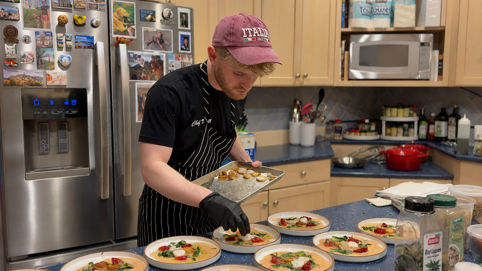A man wearing a pink cap, black gloves, and a black and white striped apron is preparing and plating bowls of soup with herbs, tomatoes, and cheese in a kitchen. The countertop has multiple bowls of soup, and the kitchen has wooden cabinets, a microwave, and a refrigerator covered with magnets and photos.