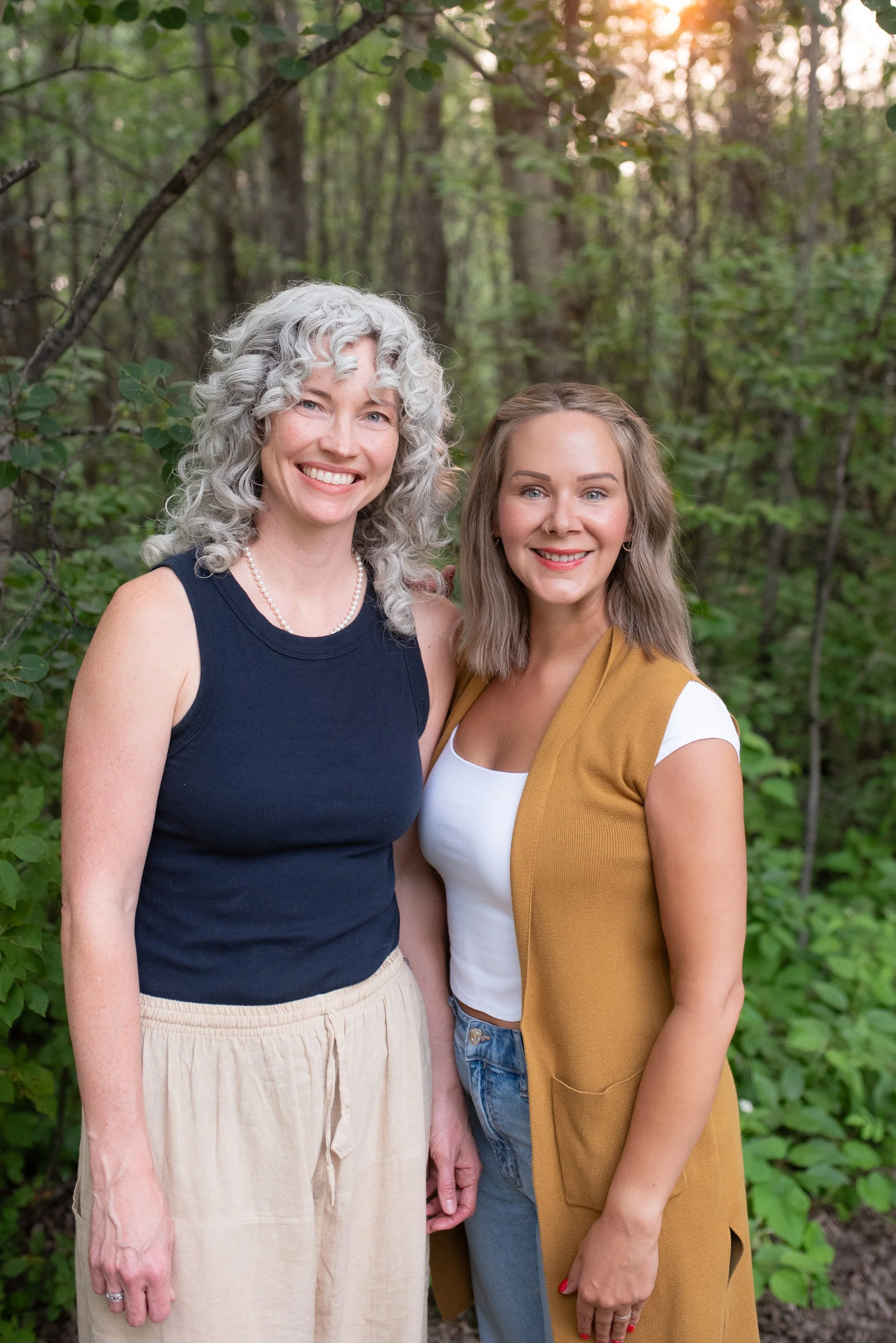 Two women standing outdoors in a wooded area, smiling at the camera. The woman on the left has curly gray hair and is wearing a black sleeveless top and beige pants. The woman on the right has wavy blonde hair and is wearing a white shirt with a mustard-colored vest. The background features green trees and sunlight filtering through the leaves.