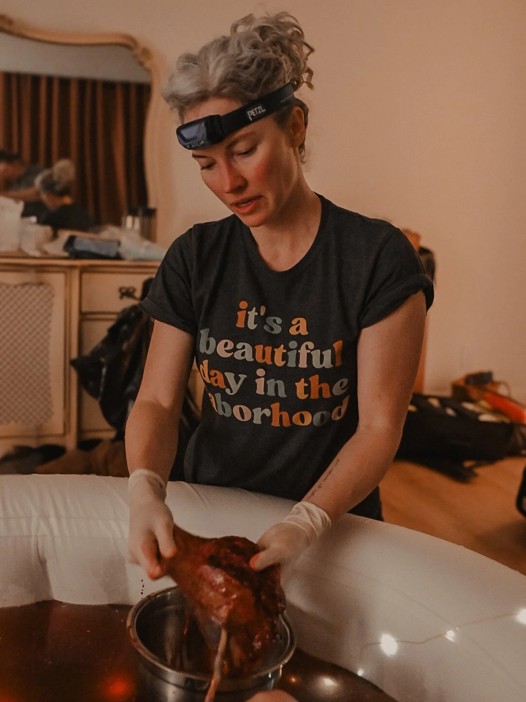 Woman wearing gloves and a headlamp, preparing or carving a large piece of meat, possibly doing a culinary or meat preparation activity, in a room with a dresser and other people in the background.
