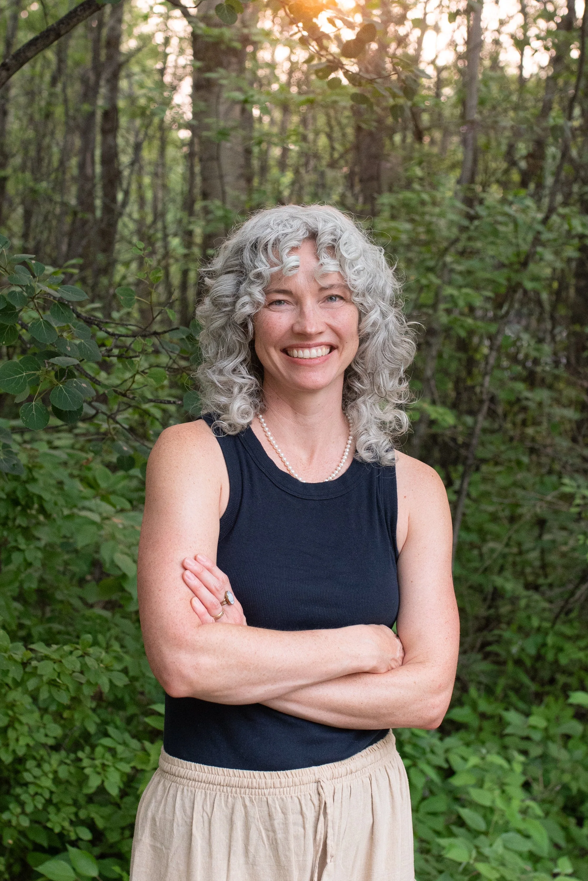 A woman with curly gray hair smiling with arms crossed, standing outdoors in a wooded area with green foliage and trees.