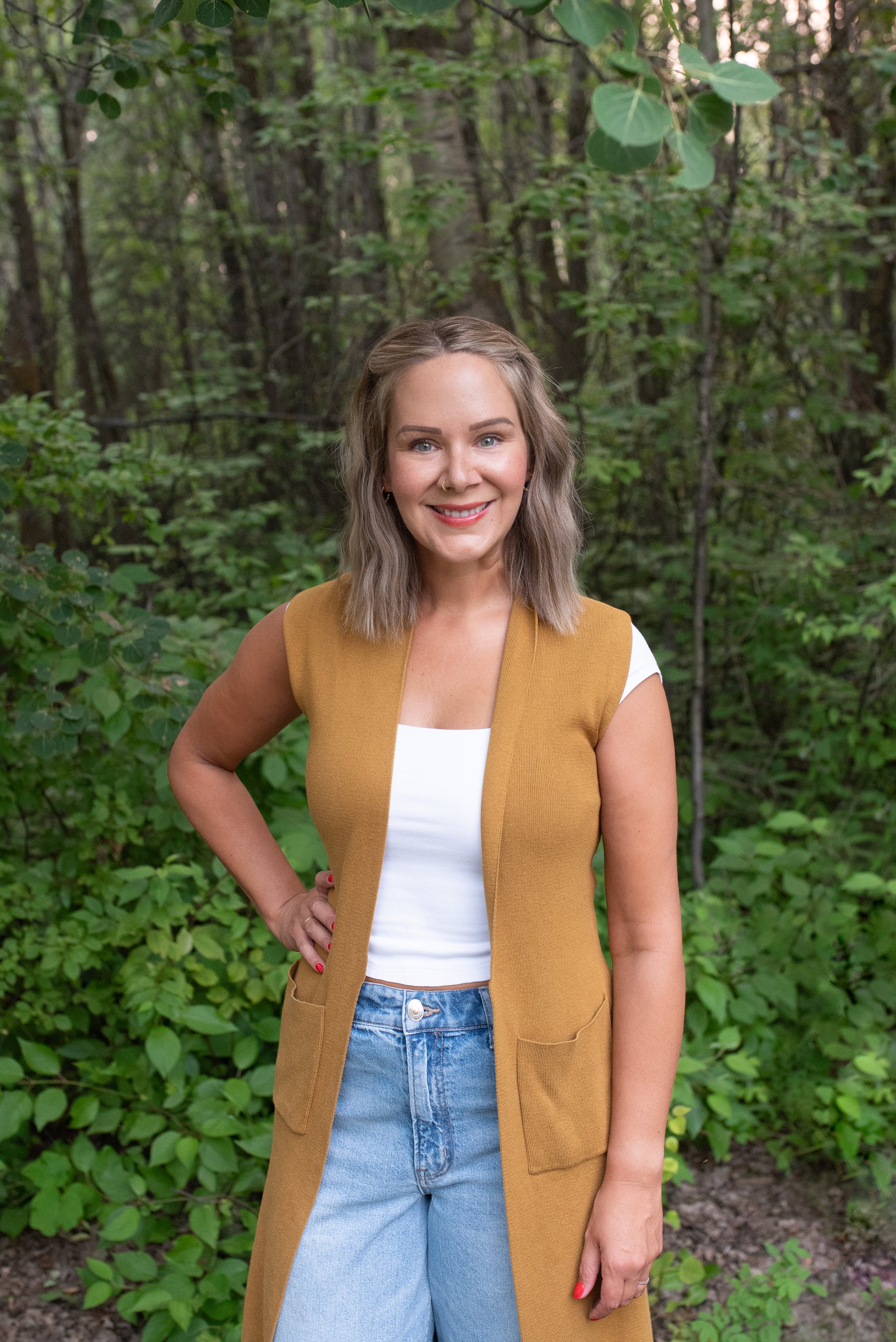 A woman with shoulder-length blonde hair, blue eyes, and a big smile, wearing a white top, a mustard yellow vest, and light blue jeans, standing outdoors in a lush green forest.